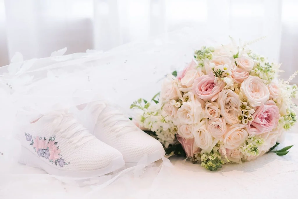 White sneakers with pastel floral embroidery next to a bouquet of pink and white roses.