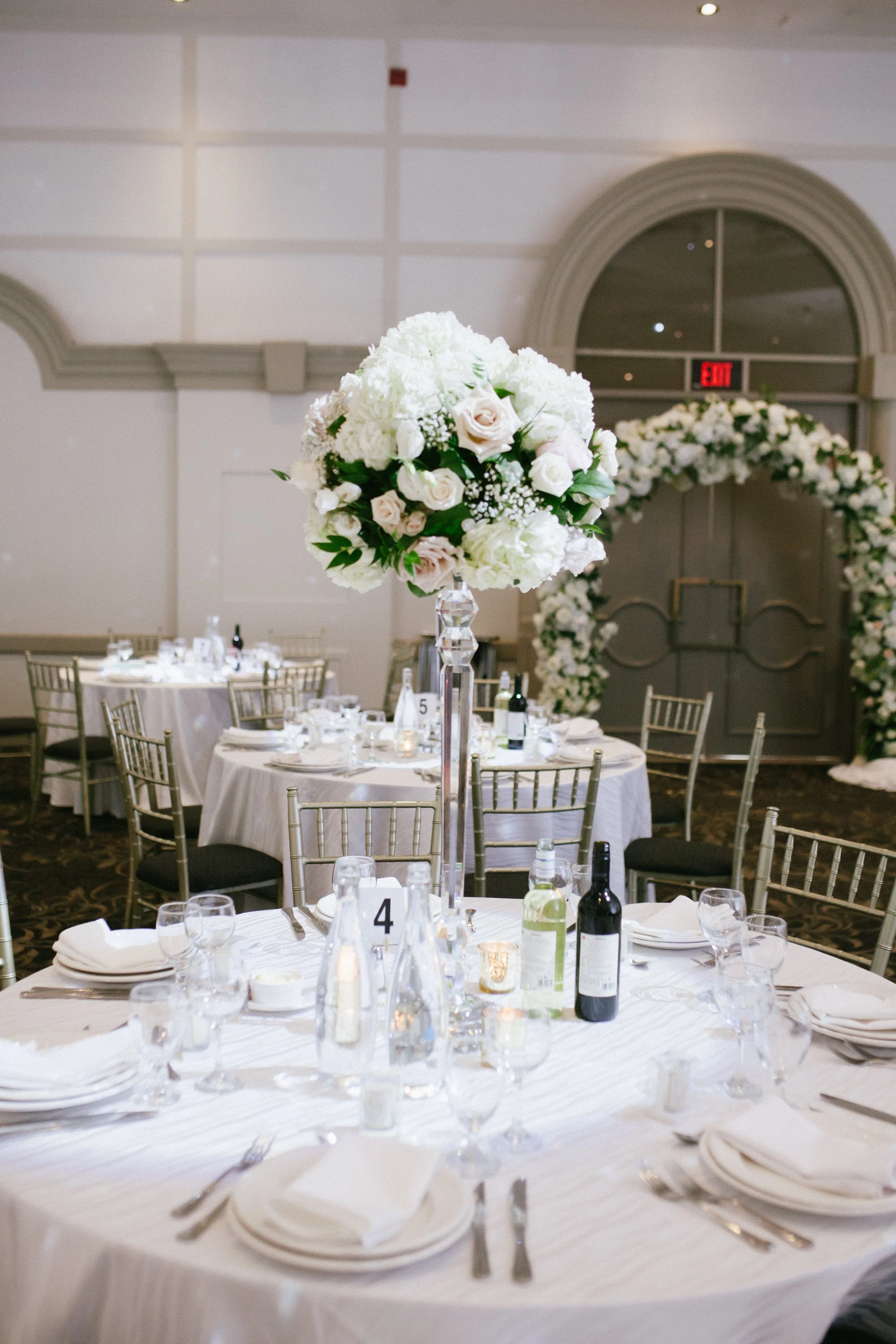 A wedding reception table with a tall floral centerpiece, set with plates, glasses, and cutlery, in a decorated banquet hall with floral archway in the background.