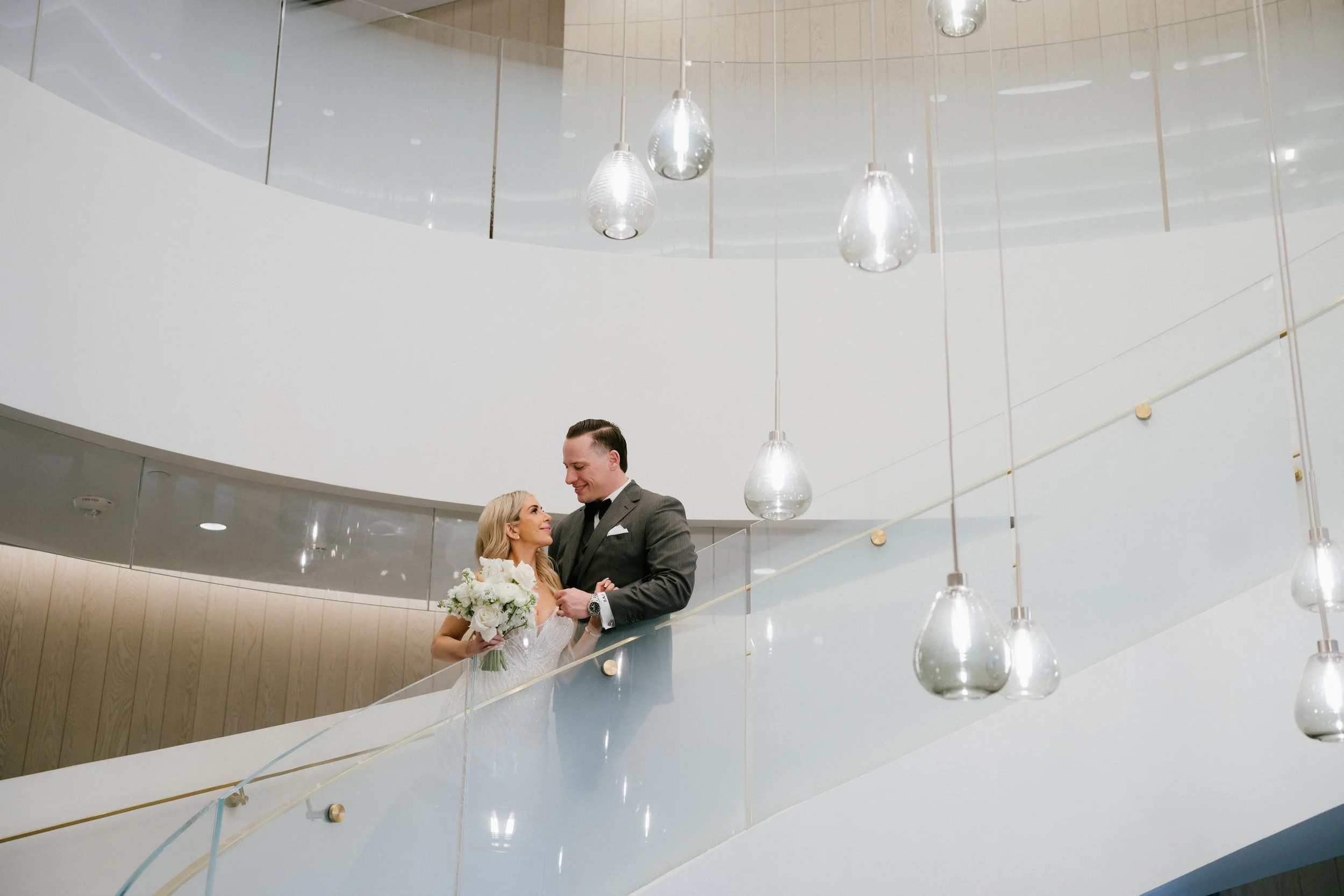 A bride and groom standing on an indoor staircase, smiling at each other. The bride holds a bouquet of white flowers, and the groom wears a dark suit with a watch on his wrist. Modern hanging lights above them, surrounded by a sleek, minimalist interior design.