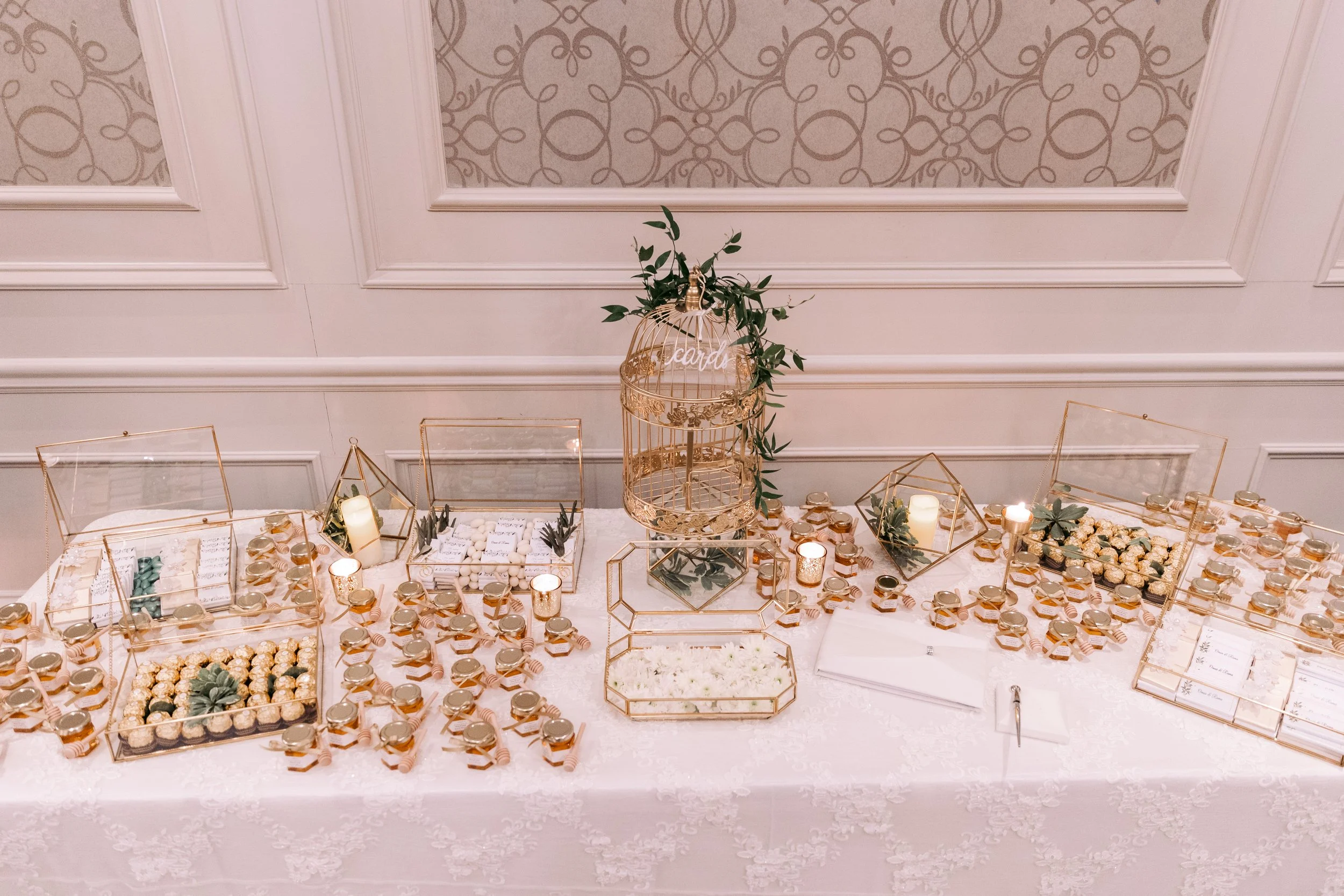 Table decorated for a wedding or party with glass boxes filled with sweets, candles, and greenery, and a birdcage in the center with a sign reading 'Cards'.