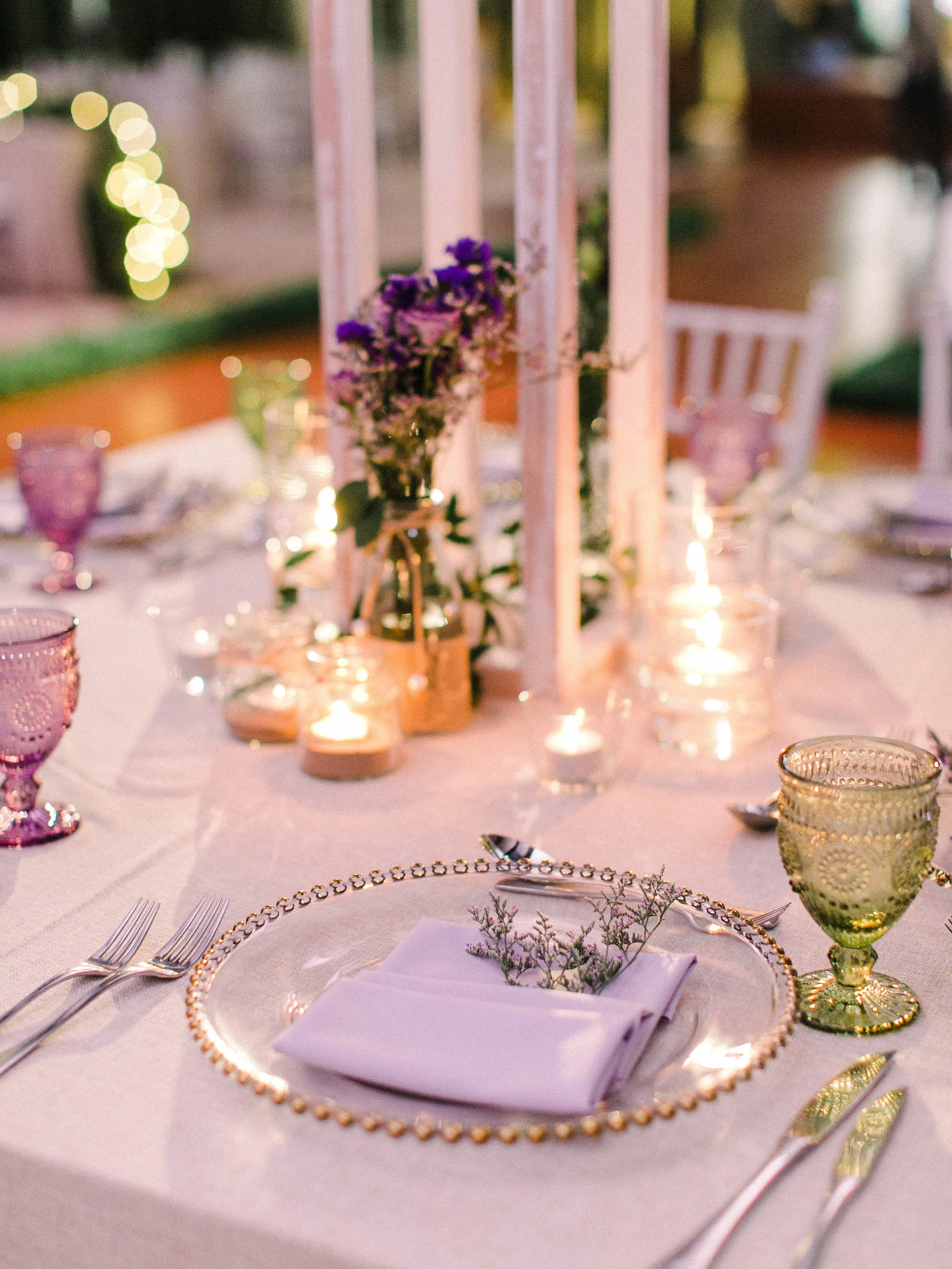 Elegant outdoor dinner table decorated with candles, flowers, and pastel-colored glassware, set for a special occasion.
