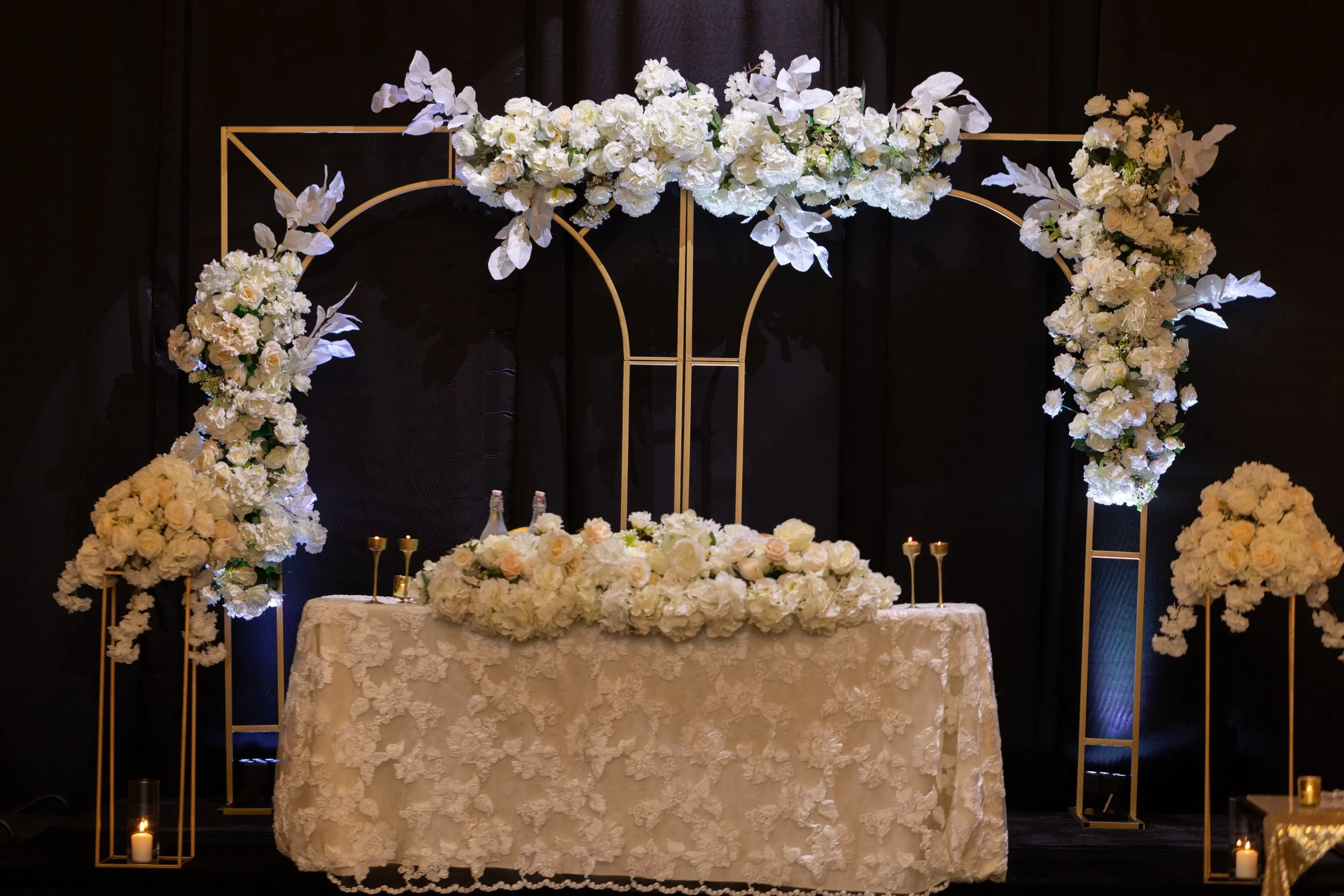 Wedding floral arch with white roses and greenery, golden stands, candles, and a lace tablecloth on a black backdrop.