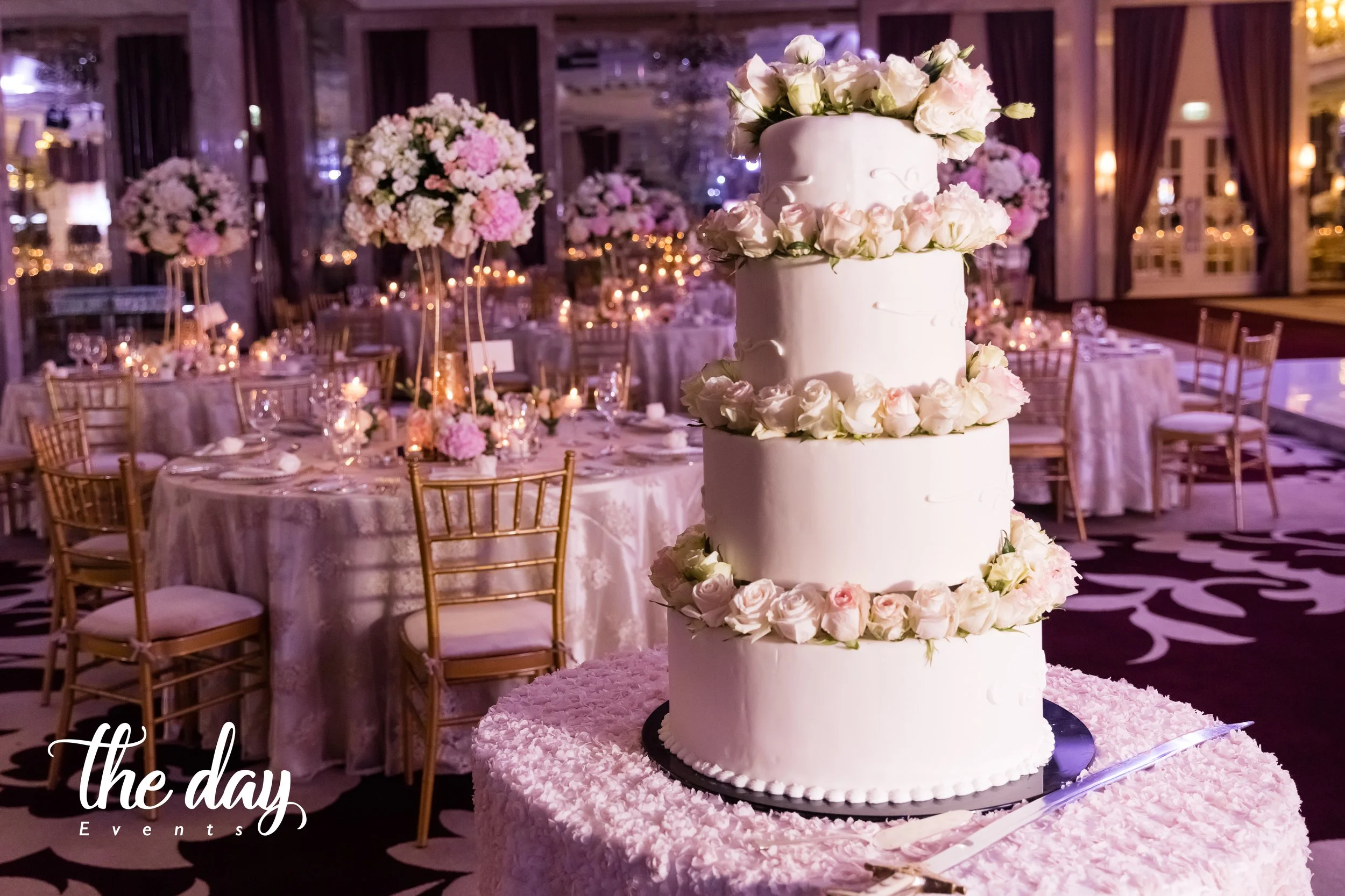 A three-tier white wedding cake decorated with pink and white roses, placed on a pink textured table in an elegant banquet hall with floral centerpieces and gold chairs.