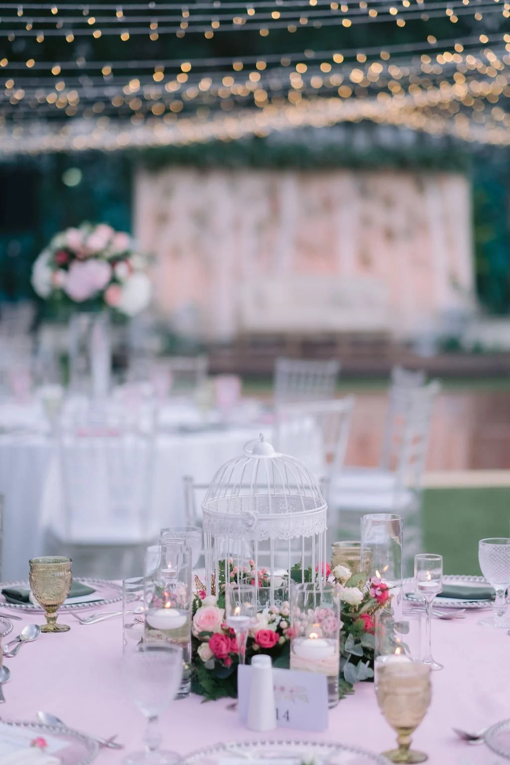 Wedding reception table with pink and white floral centerpiece, lanterns, candles, glasses, and silverware, with a blurred background of string lights and banquet hall decorations.