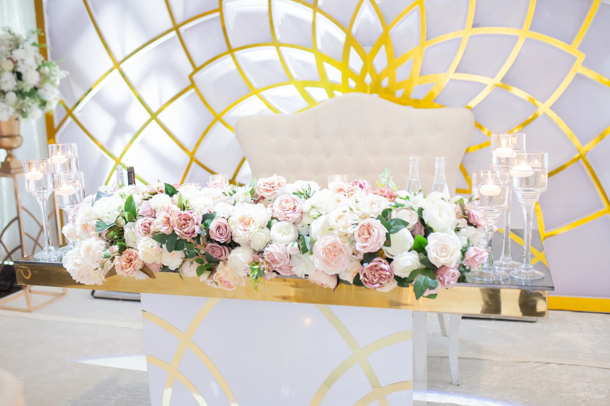 A floral arrangement of pink and white roses on a table, with glass candle holders, candles, and glass bottles. In the background, a gold geometric wall design and a white plush chair are visible.