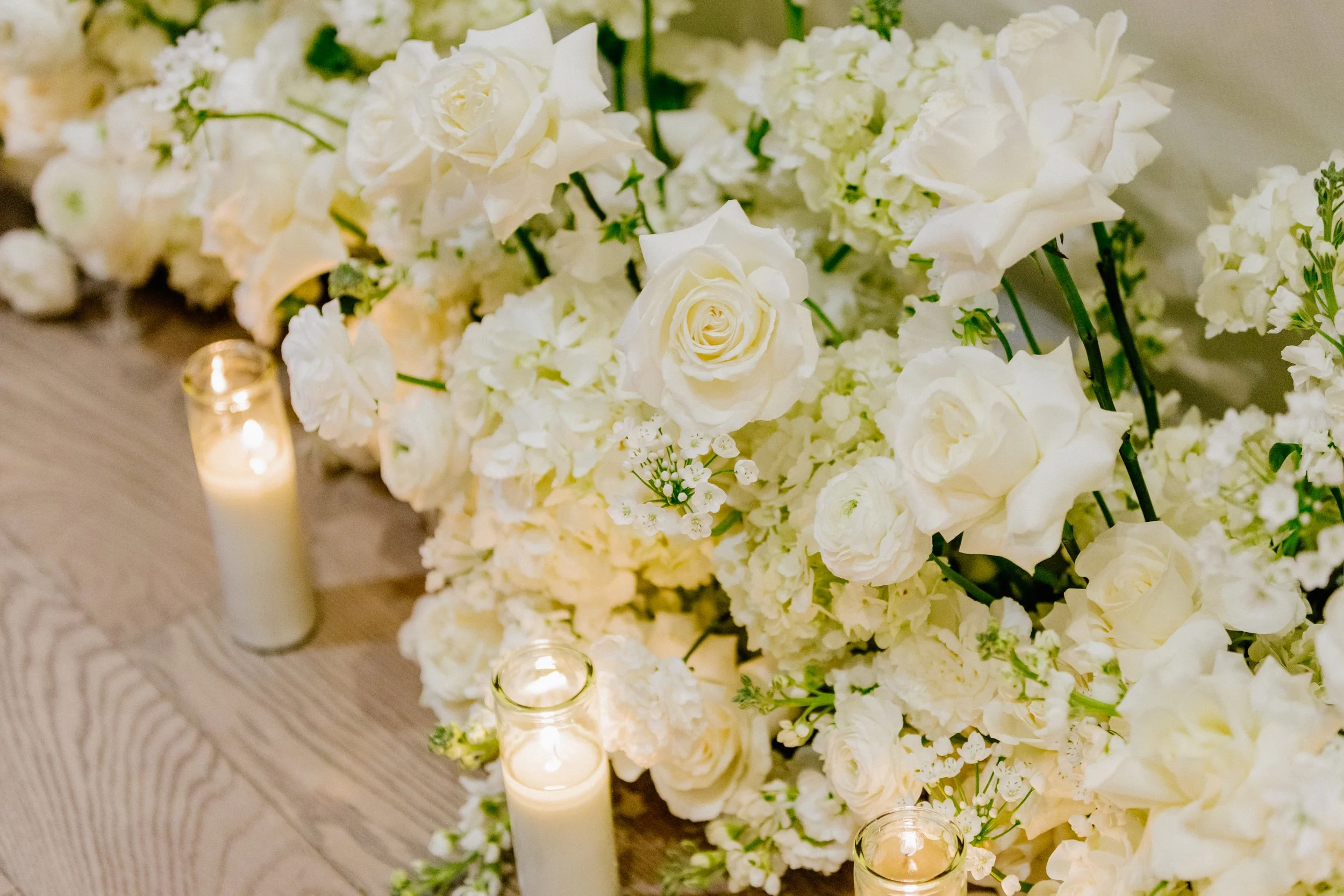 White floral arrangement with roses, hydrangeas, and small white flowers, surrounding lit candles on a wooden surface.