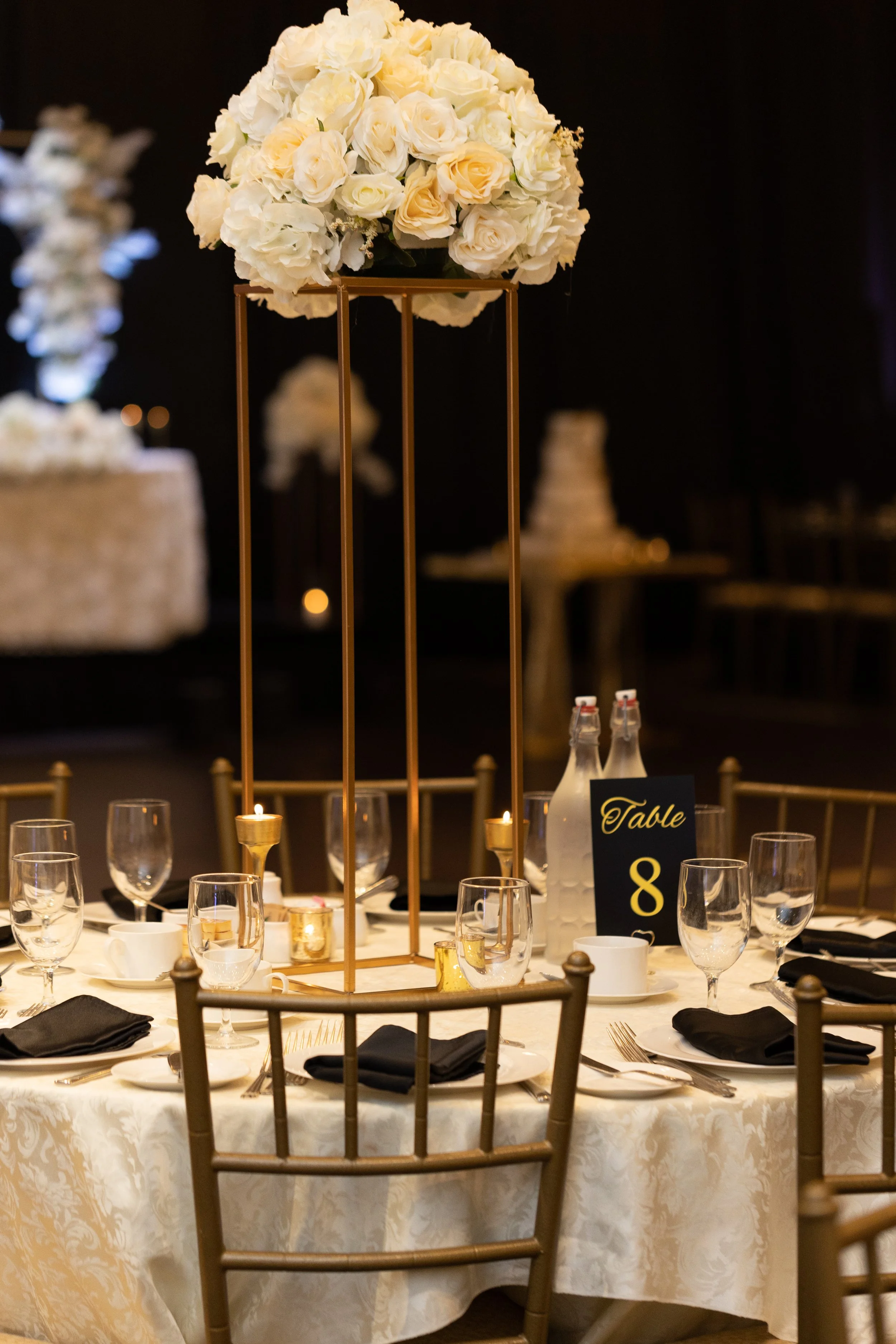 A banquet table decorated with a tall centerpiece consisting of white roses, set with wine glasses, black napkins, white plates, and gold chairs, with a black table number sign reading 'Table 8' and bottles of water.