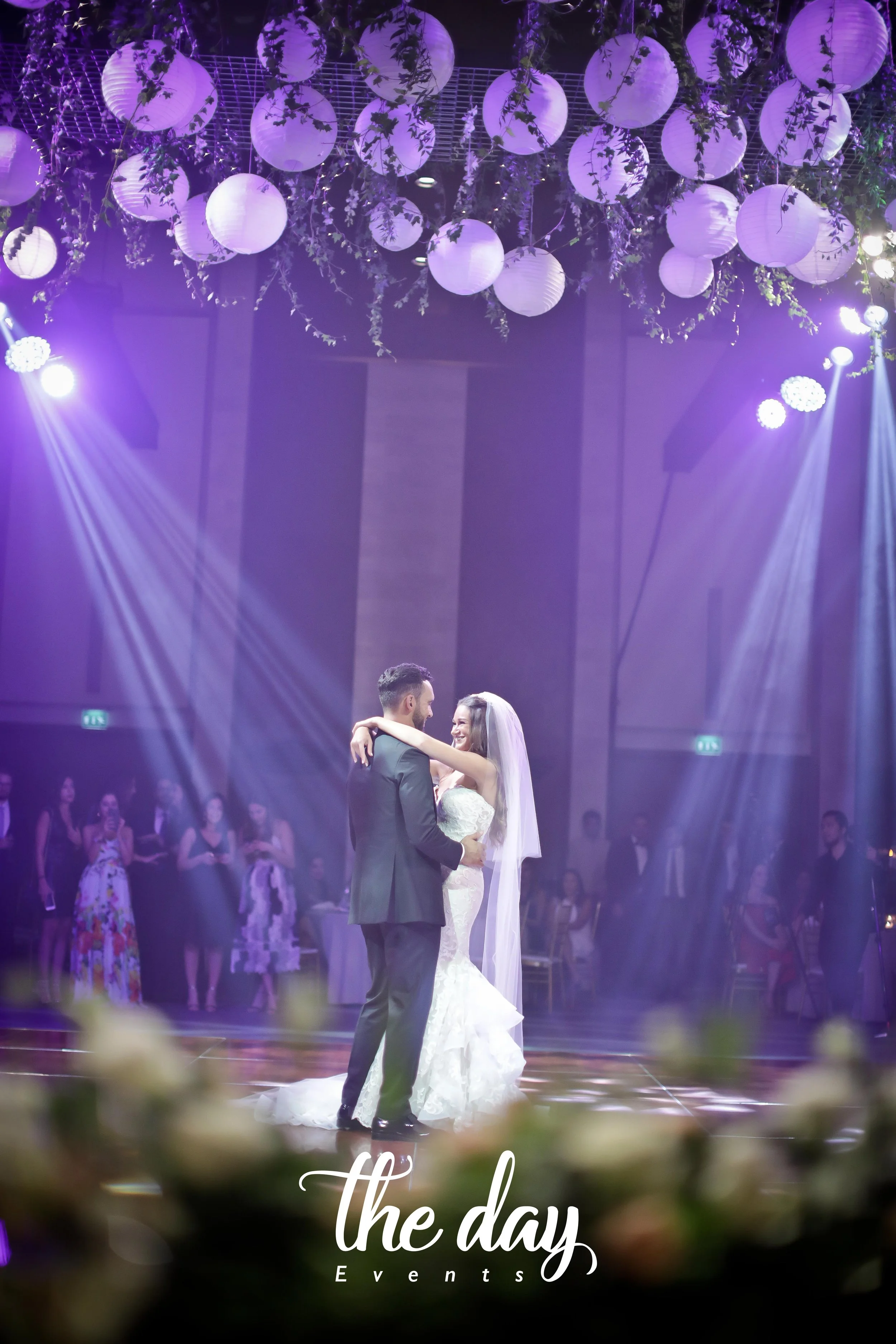 A bride and groom dance at their wedding reception with purple lighting, surrounded by guests, floral decorations, and paper lanterns hanging from the ceiling.