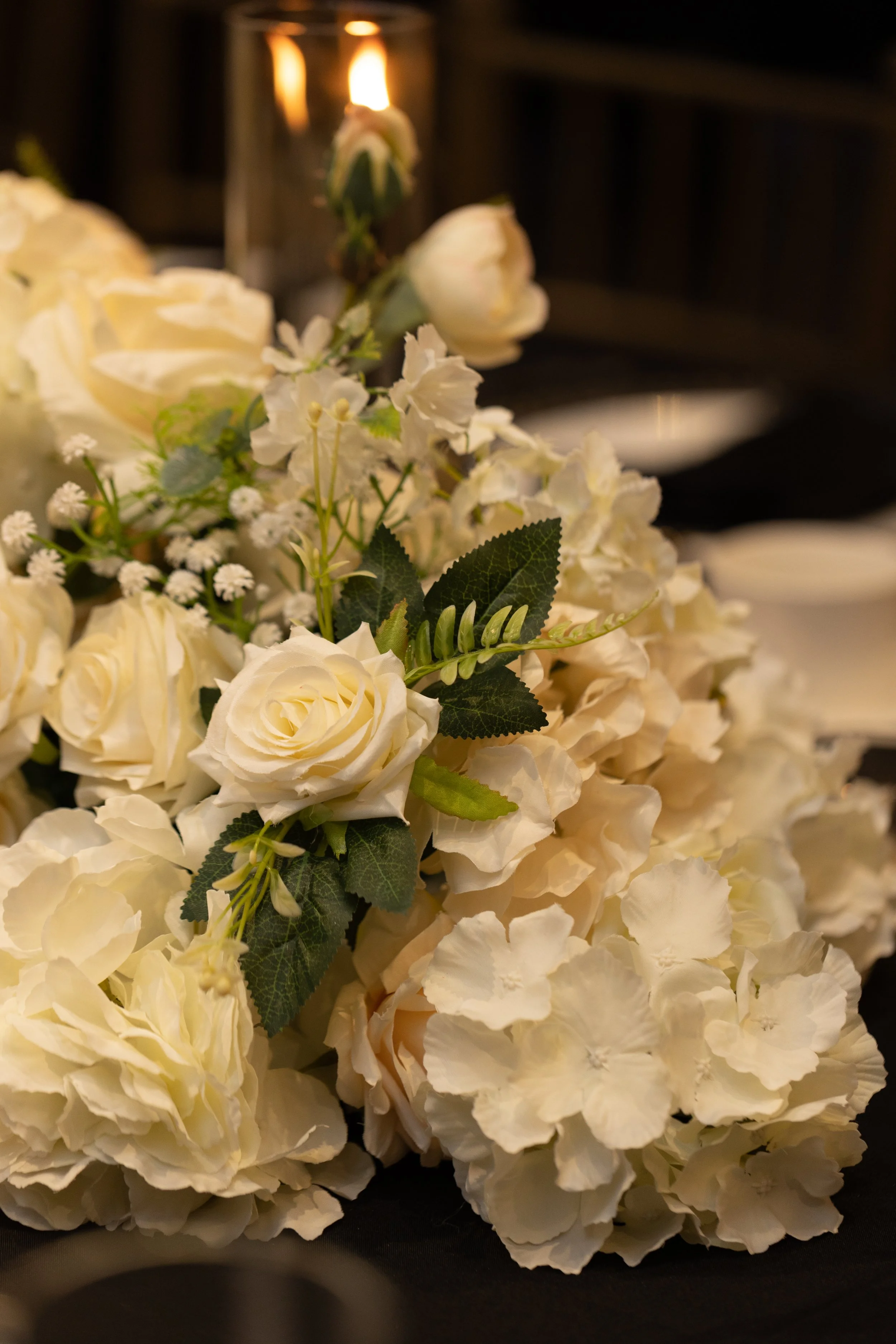 Cream-colored floral arrangement with roses, hydrangeas, and greenery on a dark table with a lit candle in the background.