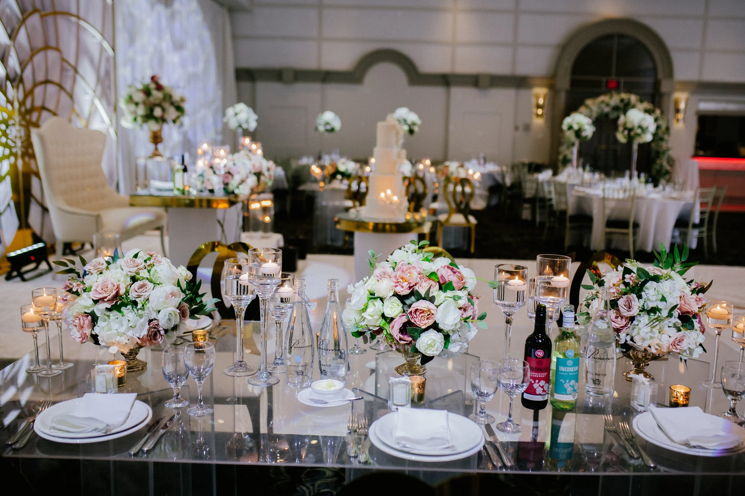 Elegant wedding reception table decorated with pink and white floral centerpieces, surrounded by candles, glasses, and table settings, with a tiered wedding cake in the background.