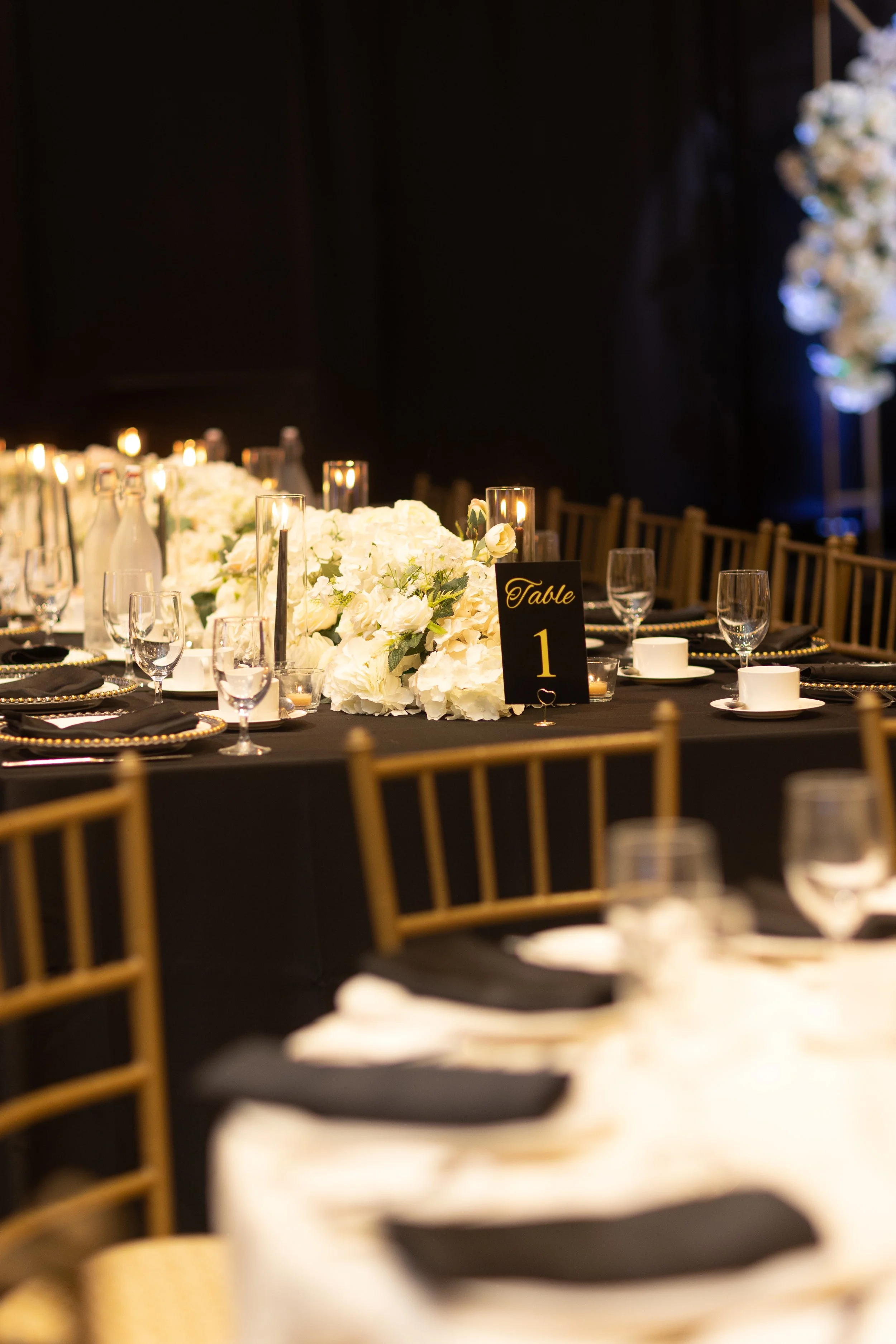 Elegant banquet table decorated with white floral centerpieces, candles, glassware, and black napkins, with a seating sign labeled 'Table 1' in black and gold.