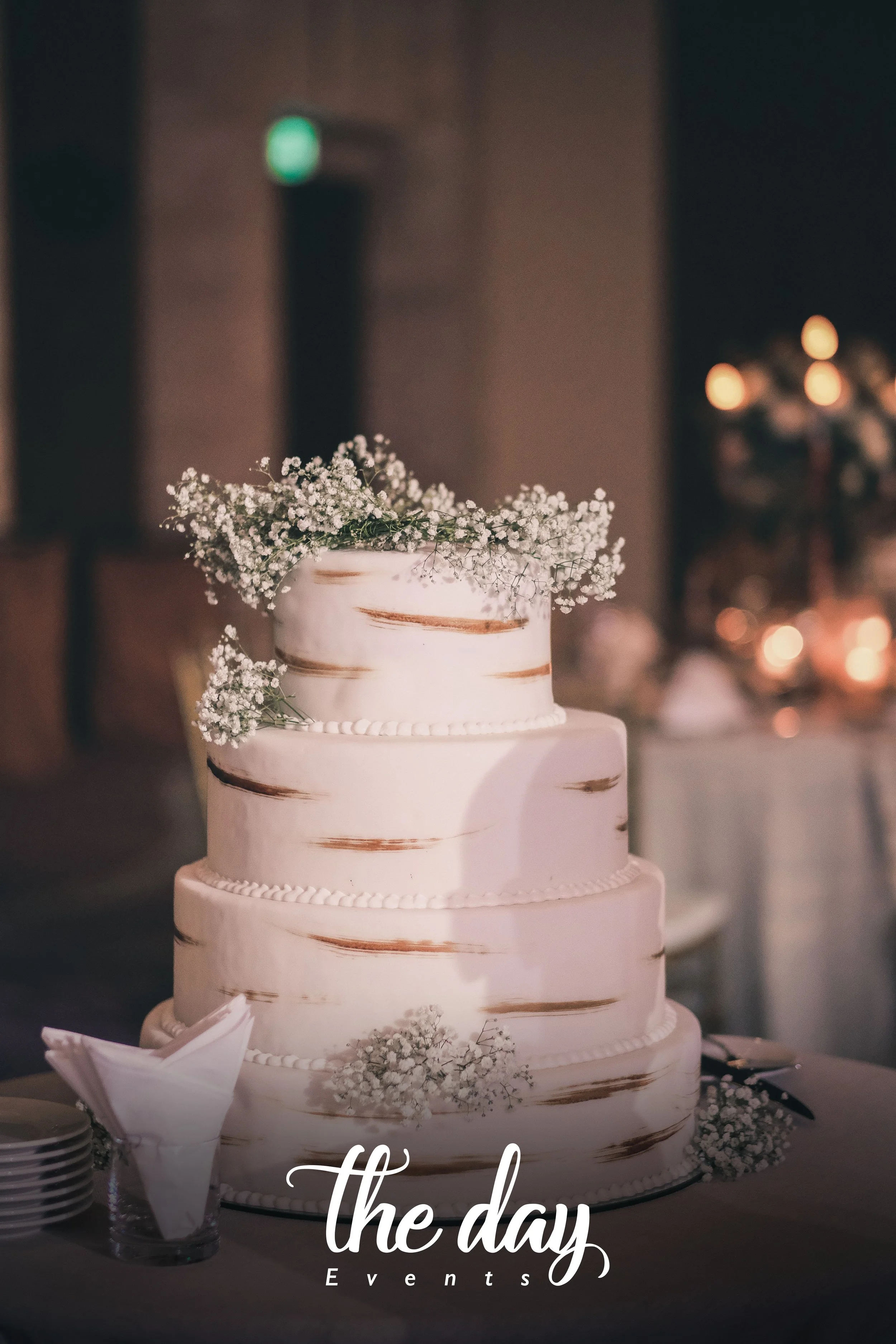 A three-tier wedding cake decorated with white and brown patterns, topped with small white flowers, on a table with white napkins and plates, with a blurred background of candles and warm lighting.