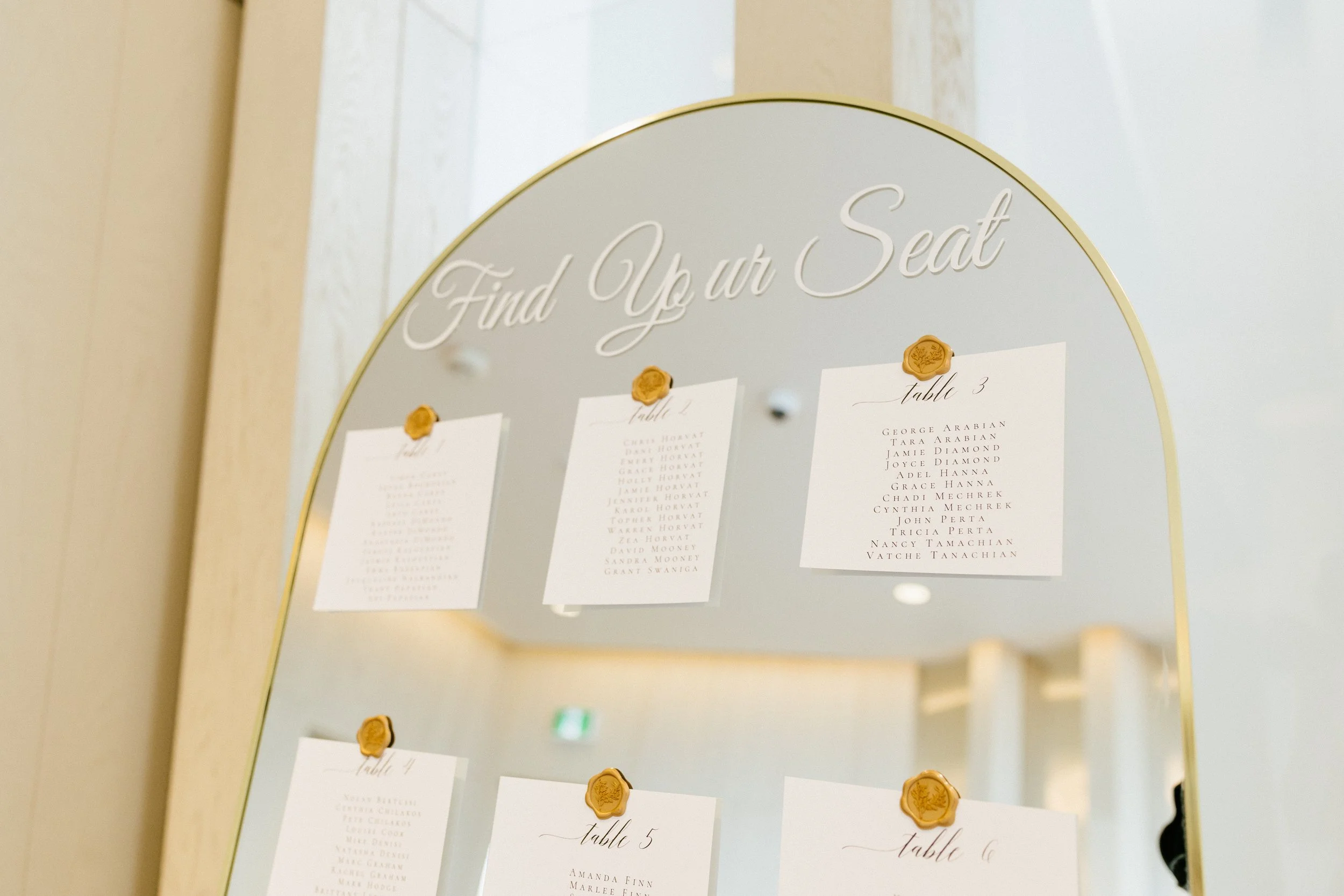 Mirror with gold accents displaying tables and guest names for a wedding, with 'Find Your Seat' written at the top.