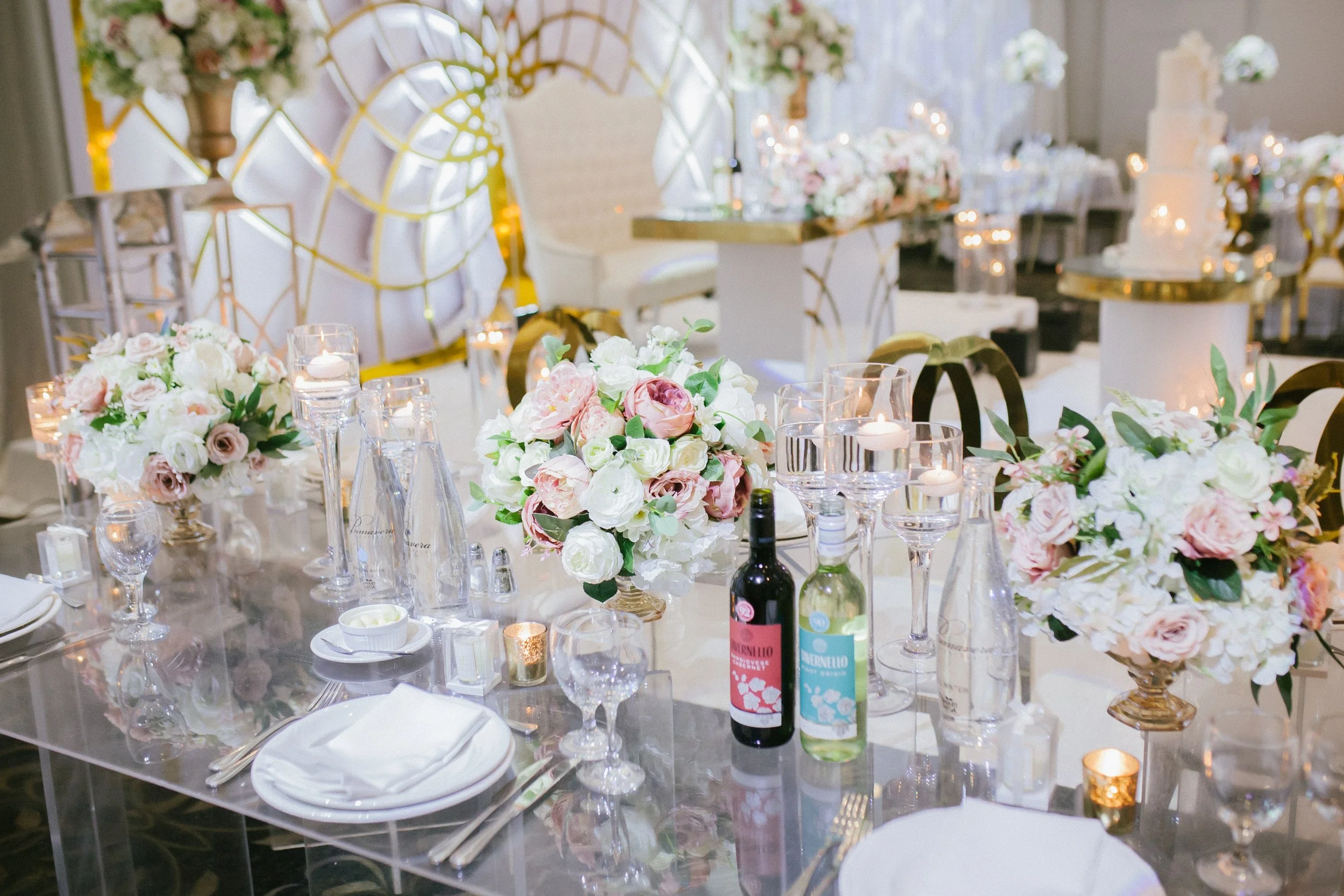 Decorated banquet table with flower centerpieces, candles, glassware, and bottles of wine, set in an elegant event hall.