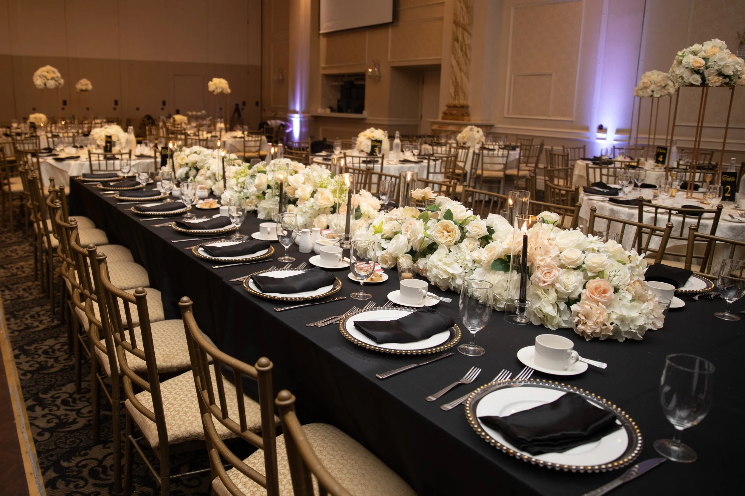 Elegant banquet hall with a long table covered in black tablecloth, decorated with white floral arrangements and candles, set for a formal event with black and gold-rimmed plates, black napkins, and clear glassware.