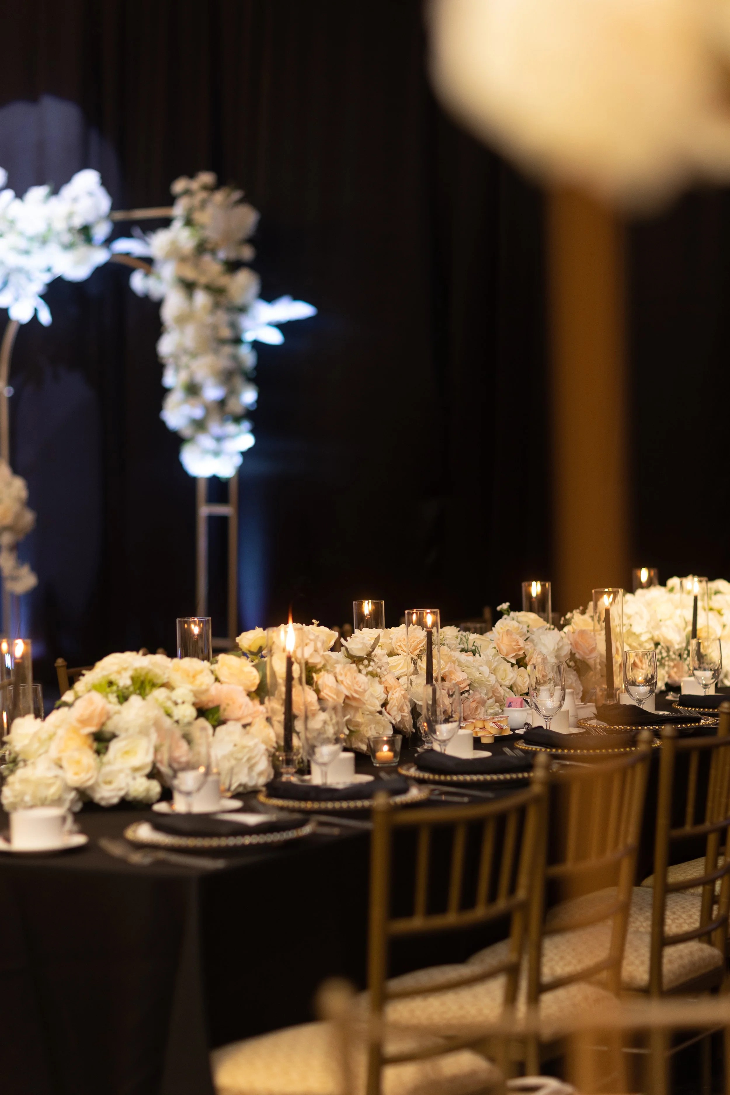 Elegant banquet table with black tablecloth, white floral centerpieces, gold chairs, and candlelit decorations at a wedding or formal event.
