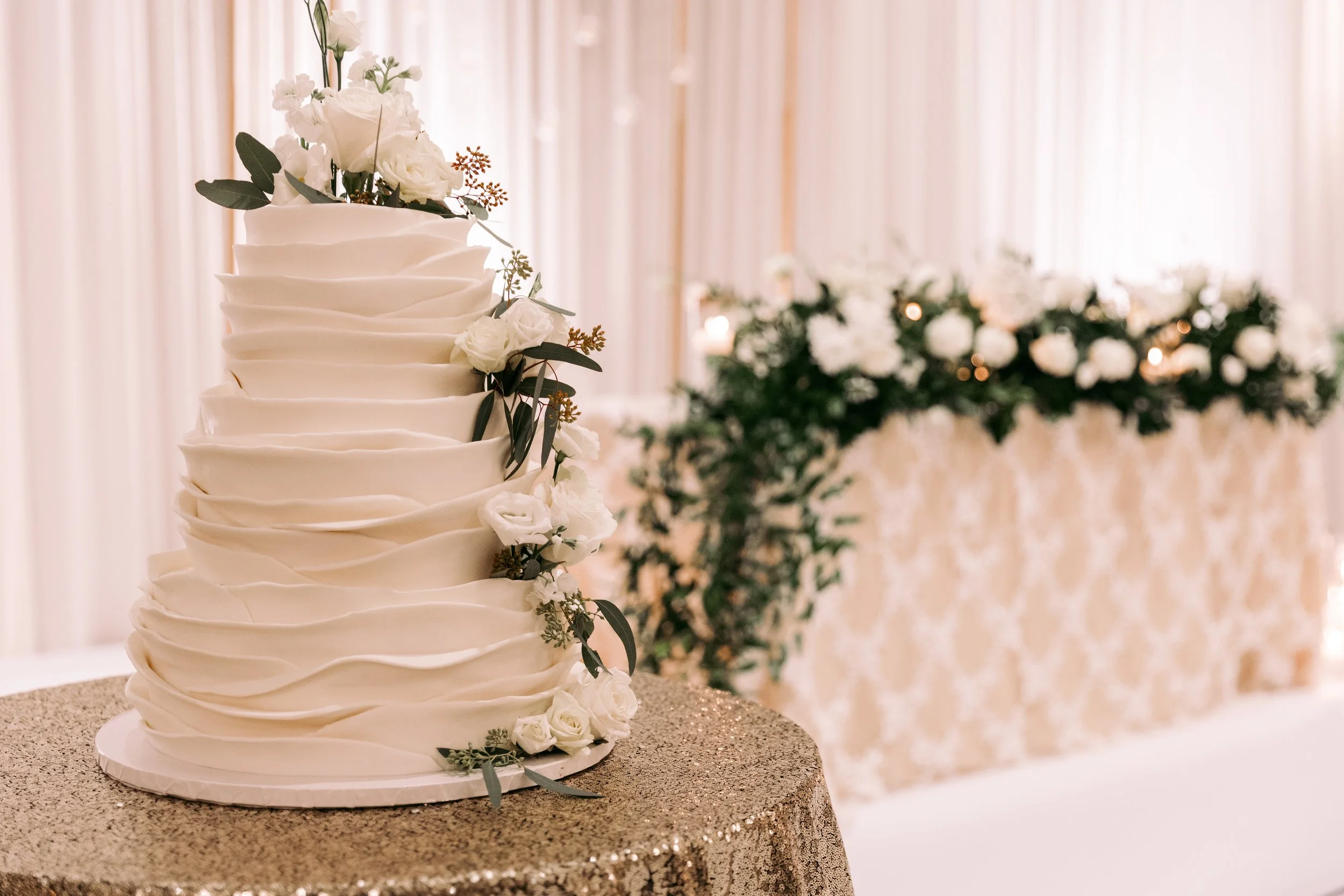 White wedding cake with a wavy textured design, decorated with white flowers and greenery, placed on a glittery gold tablecloth at a wedding reception.