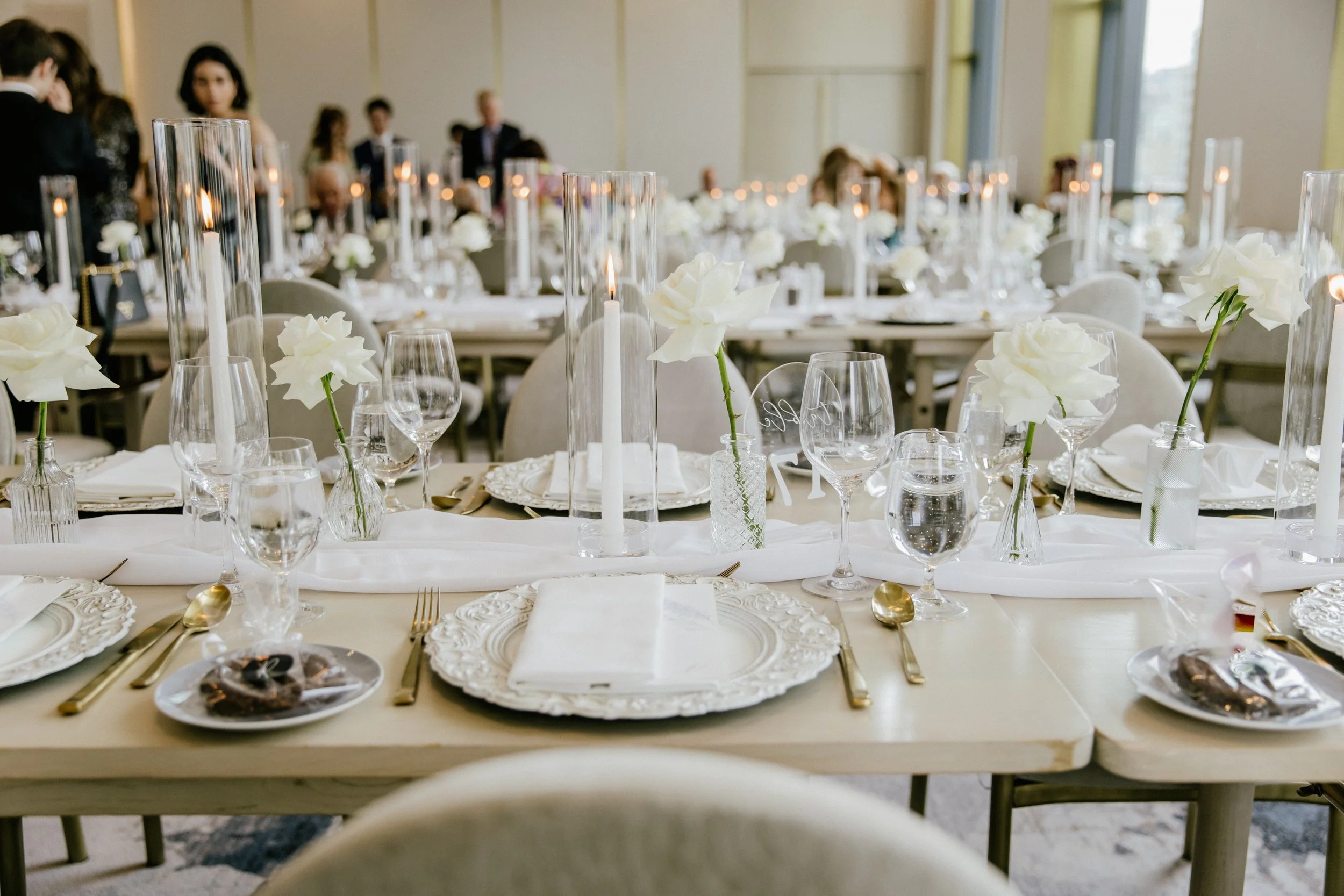 Elegant wedding reception table decorated with white flowers in vases, tall glass candle holders with candles, and gold utensils, set with ornate white plates and linen napkins. Guests are visible in the background in a well-lit room.