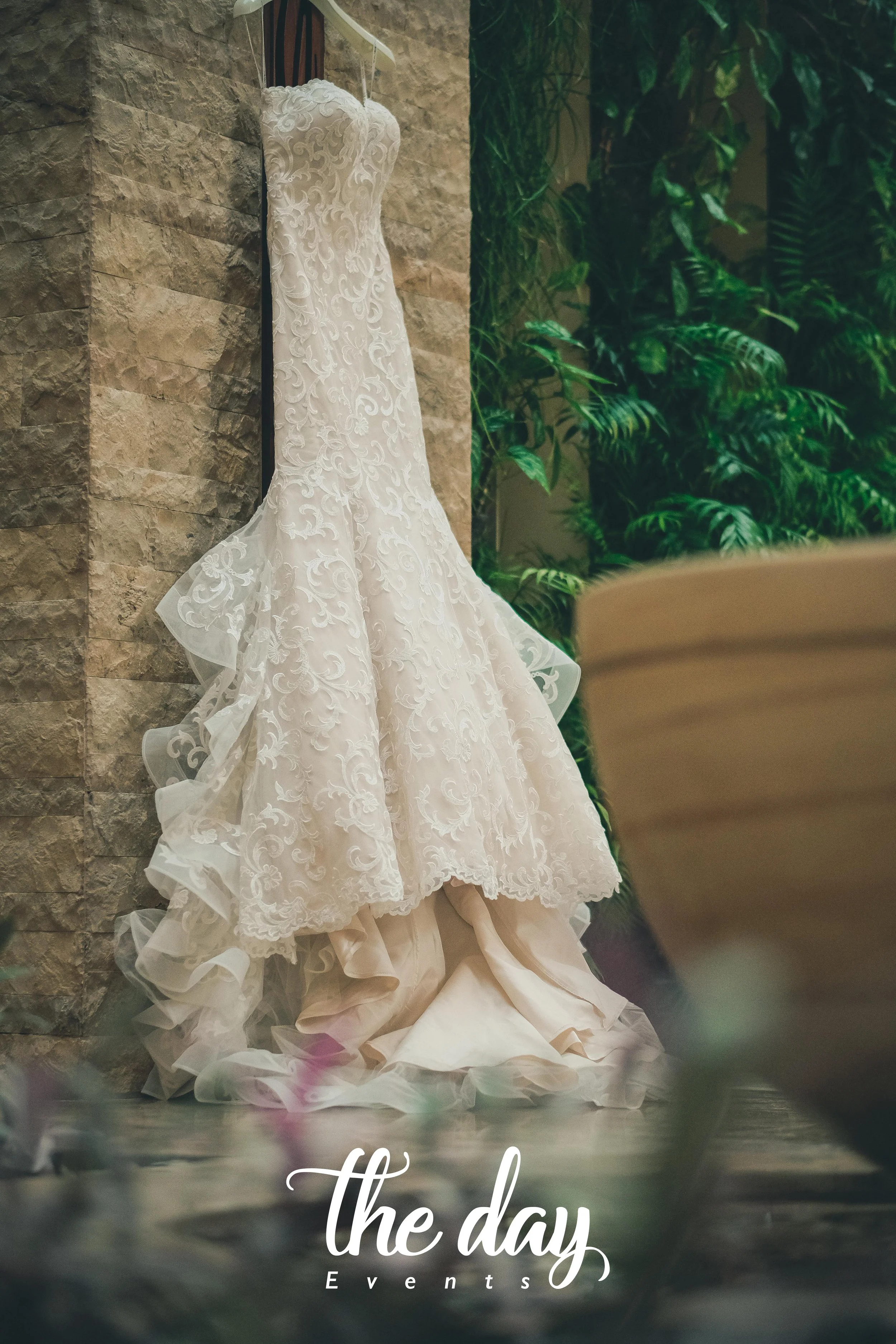 A white wedding dress with intricate lace details hangs on a hanger against a stone wall, with lush green plants in the background. Text at the bottom reads 'the day Events'.