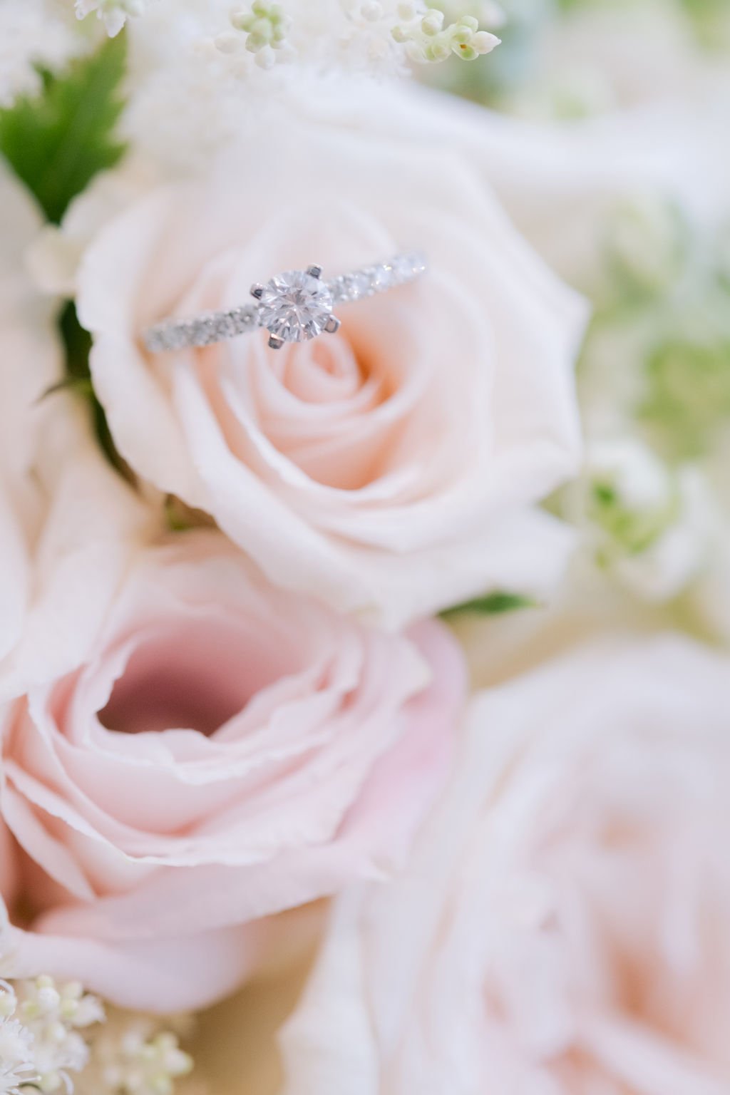 A diamond engagement ring with a central round diamond is placed on a pale pink rose in a bouquet of soft pink and white roses and green foliage.