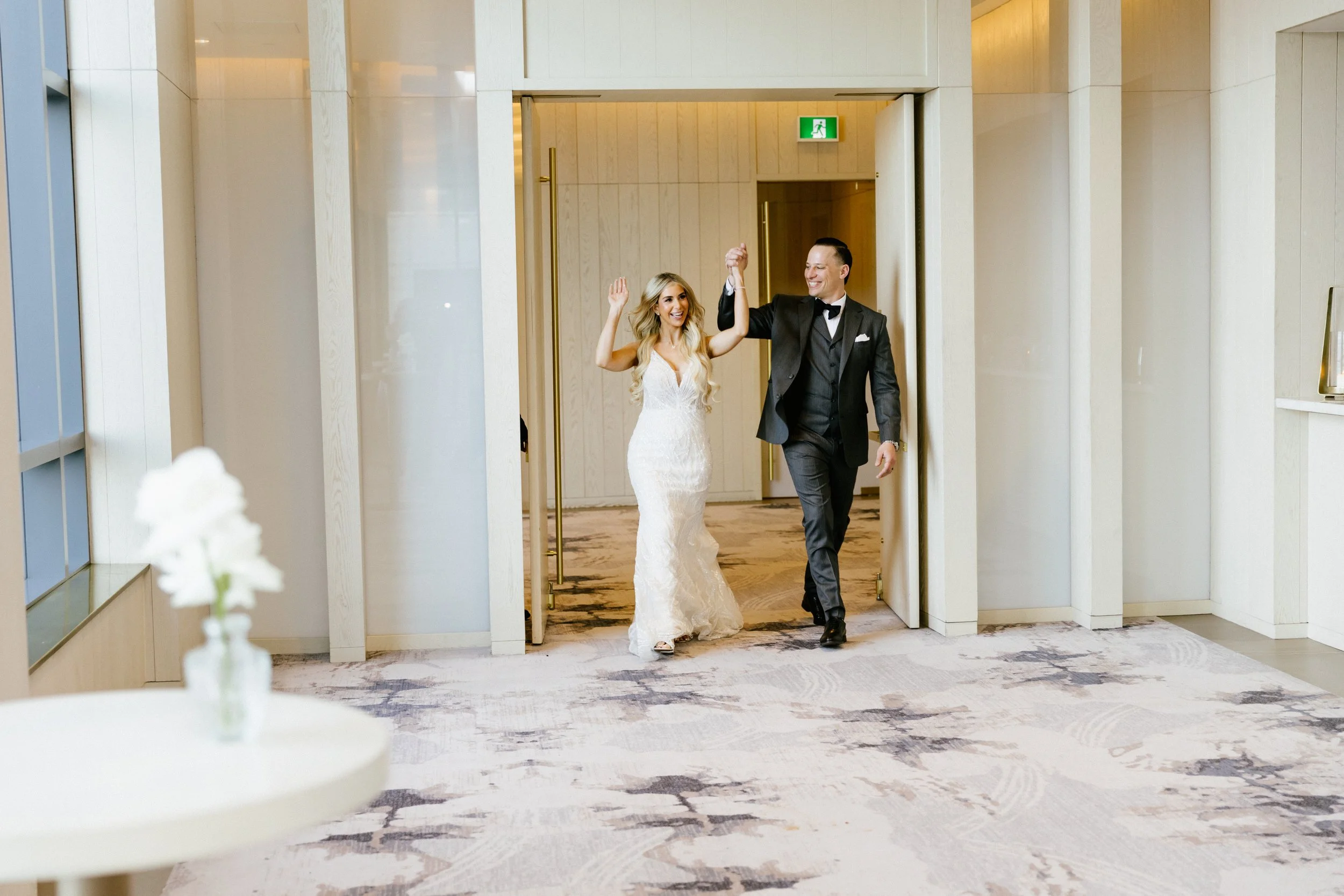 A bride and groom entering a room, celebrating and holding hands, with the bride in a white wedding dress and the groom in a black tuxedo.