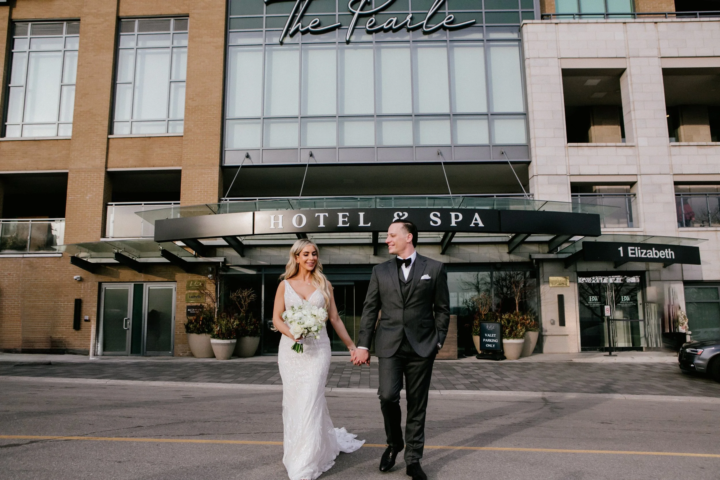 A bride and groom walking hand in hand outside a hotel and spa, smiling at each other. The bride is in a wedding dress holding a bouquet, and the groom is in a black tuxedo.