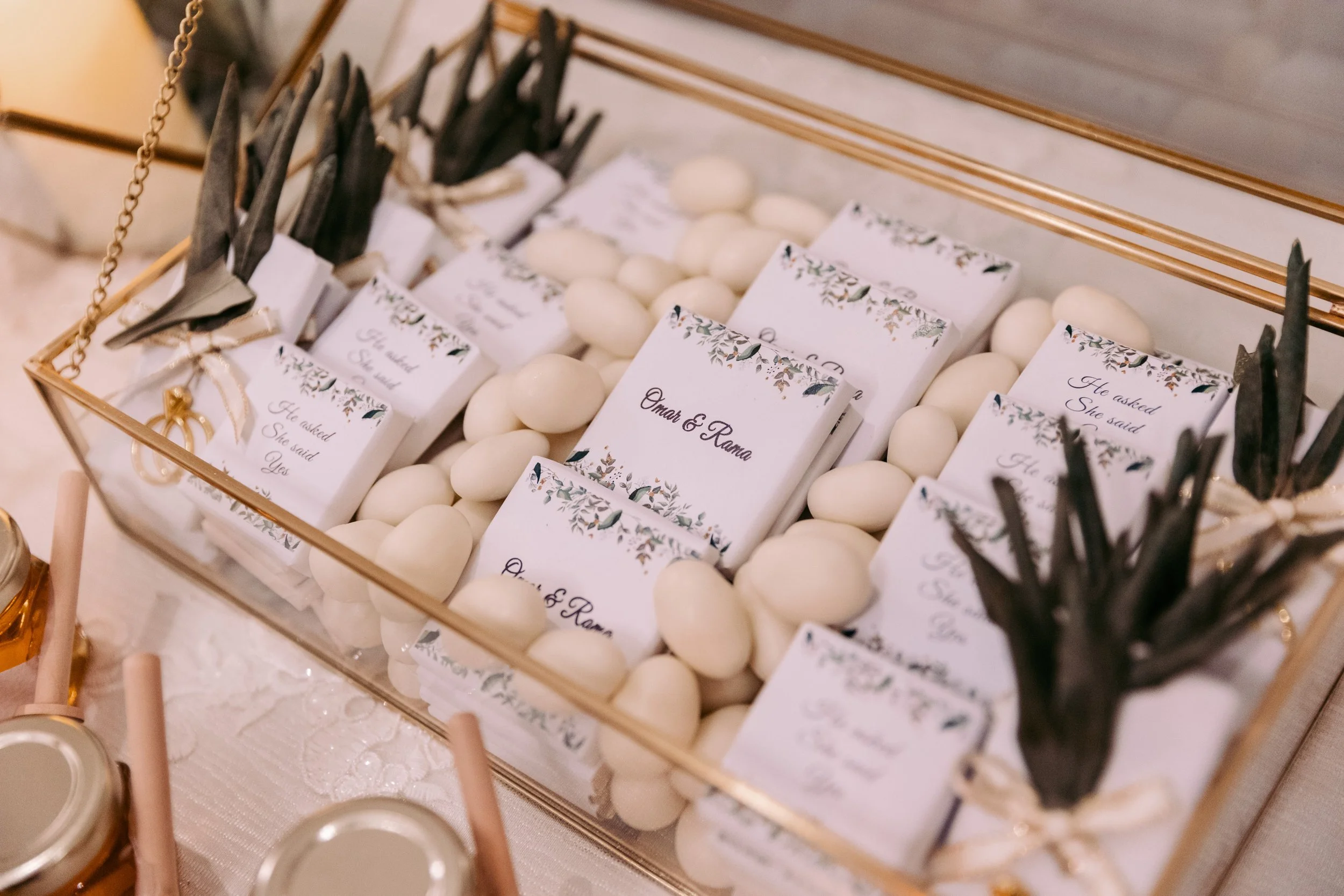 A decorative wedding favor tray with small favor boxes labeled 'Omar & Rano,' each surrounded by white candy-coated almonds, with some black feathers tied with a ribbon for decoration.