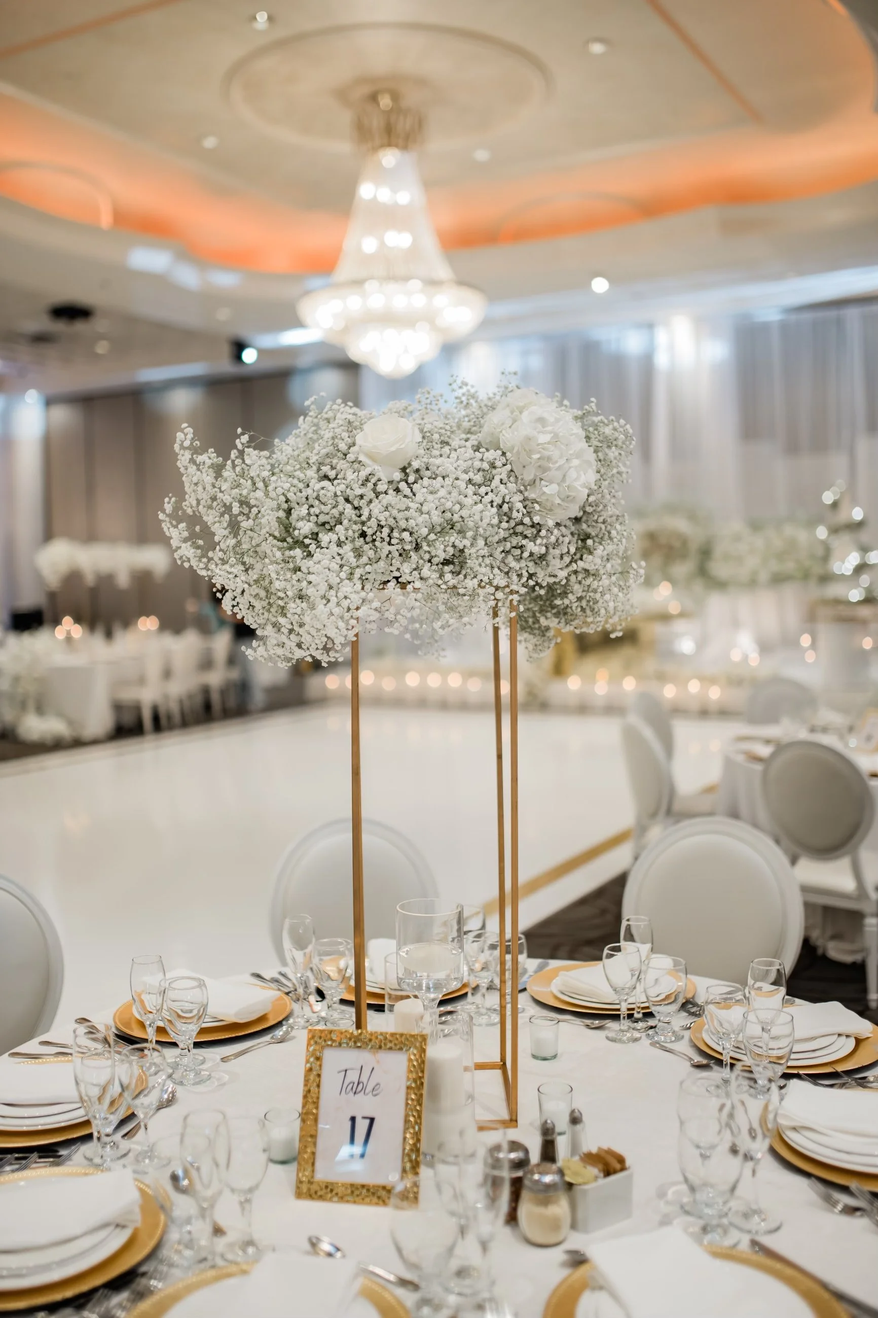 Elegant wedding reception table with a large floral centerpiece of white roses and baby's breath on a gold stand, surrounded by white and gold table settings, glassware, and candles in a decorated venue with chandeliers and draped curtains.