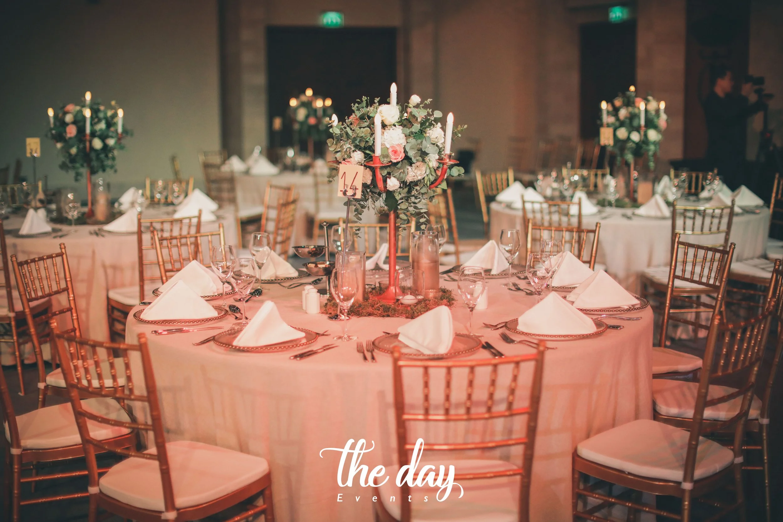 A decorated round banquet table at an event, with white tablecloth, neatly folded white napkins, wine glasses, and silverware. The centerpiece is a floral arrangement with roses, eucalyptus, and four red candles. Several similar tables are visible in