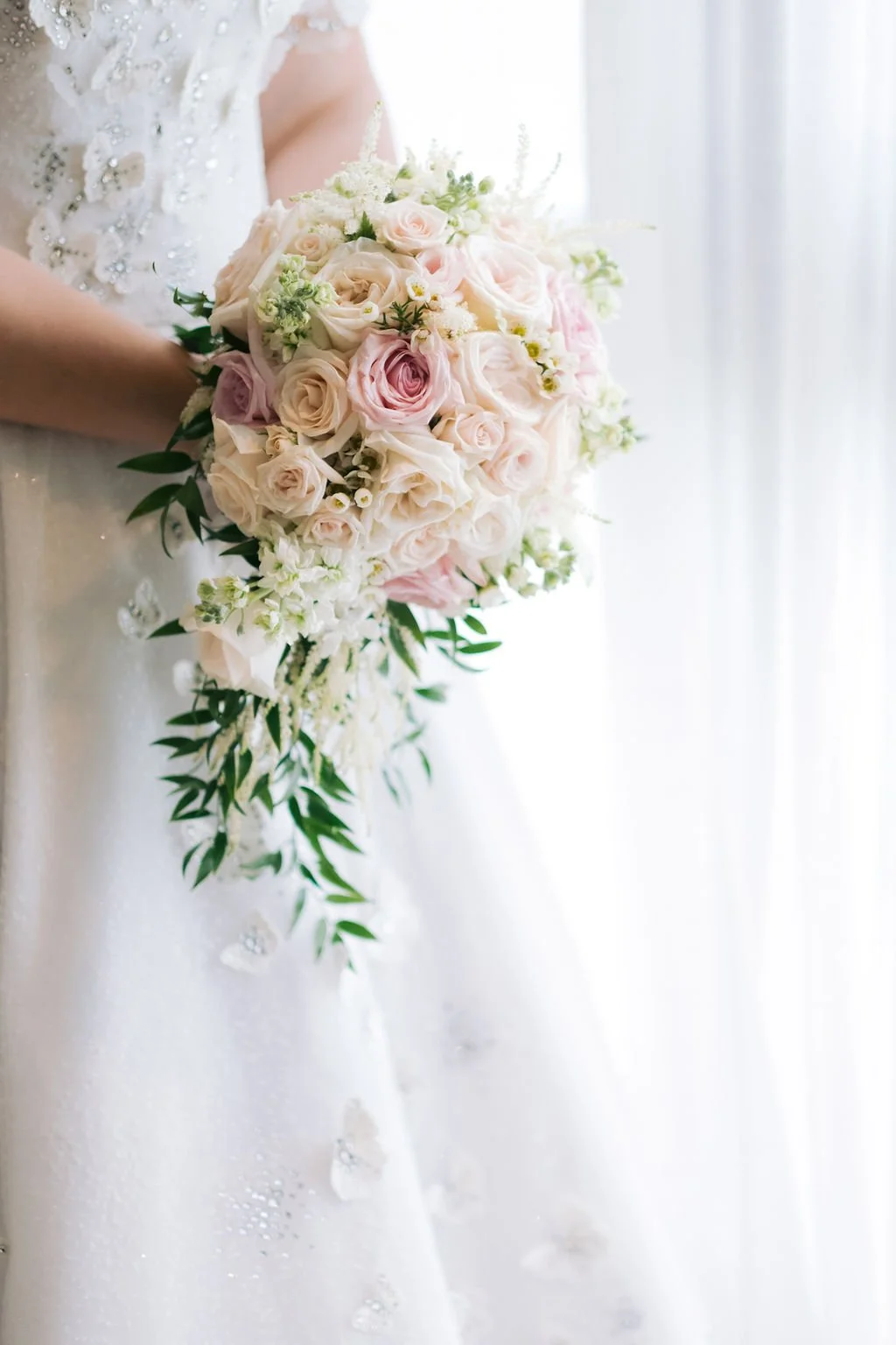 A bride holding a cascading bouquet of pink and white roses with greenery, in front of a white curtain.