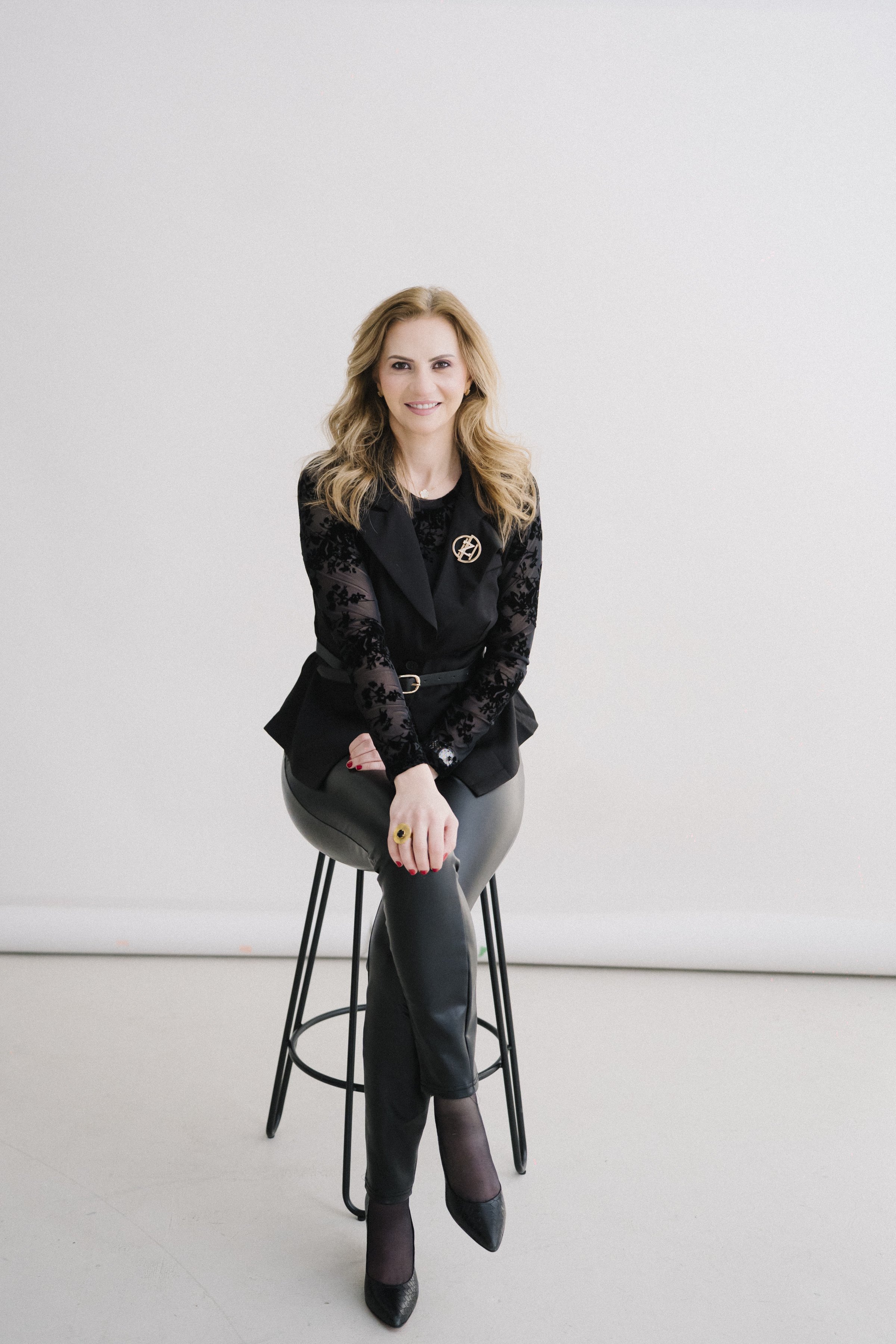 A woman with blonde hair in black leather pants and a black lace top, sitting on a stool against a plain white background.