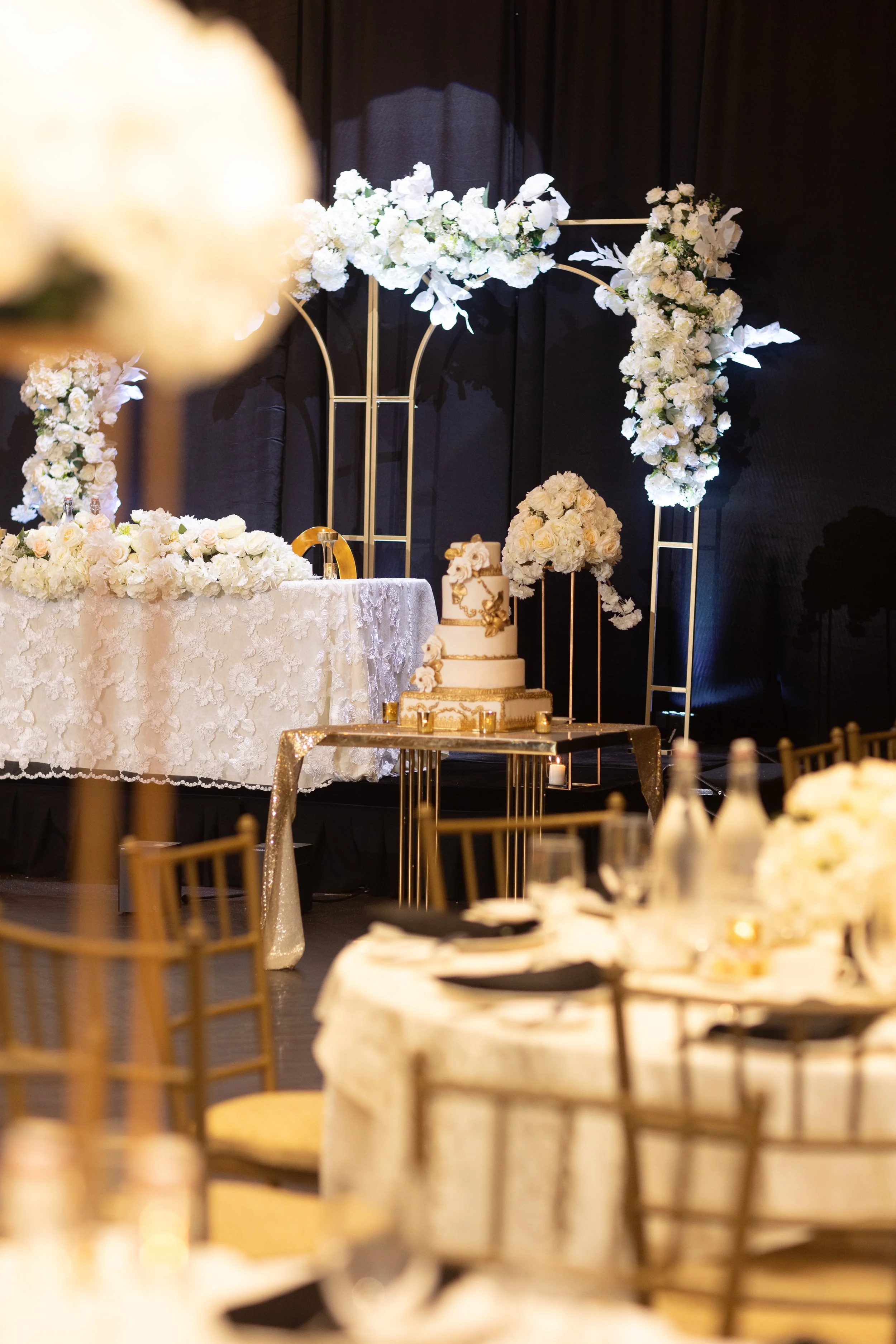 Elegant wedding reception setup with a decorated cake on a gold and glass table, surrounded by gold chairs, and floral arrangements with white flowers and green foliage on a black backdrop.