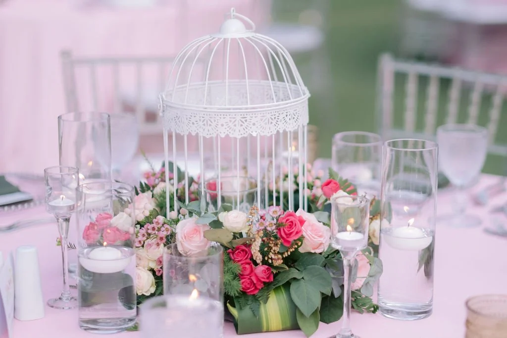 A table centerpiece with an empty white birdcage surrounded by pink, white, and red roses and greenery, with water candles in glass holders.