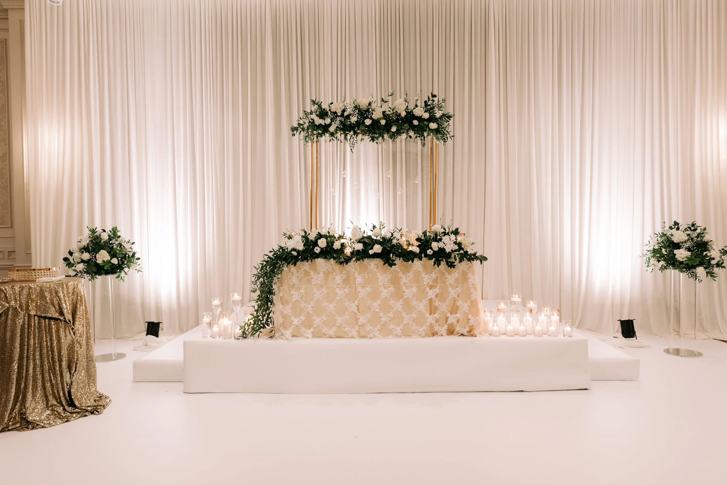 Elegant wedding altar with floral arrangements and candles, draped white curtains in the background, and a gold sequin table to the side.
