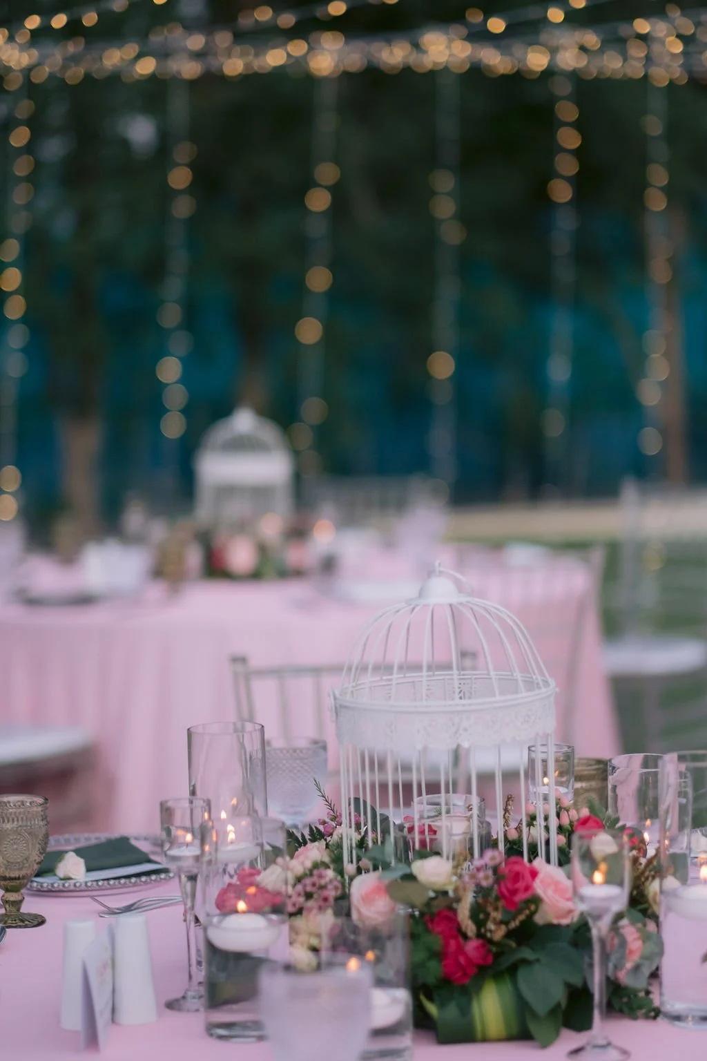 Decorated outdoor event table with pink tablecloth, floral centerpiece, birdcage, candles, glasses, and cutlery, under string lights with trees in the background