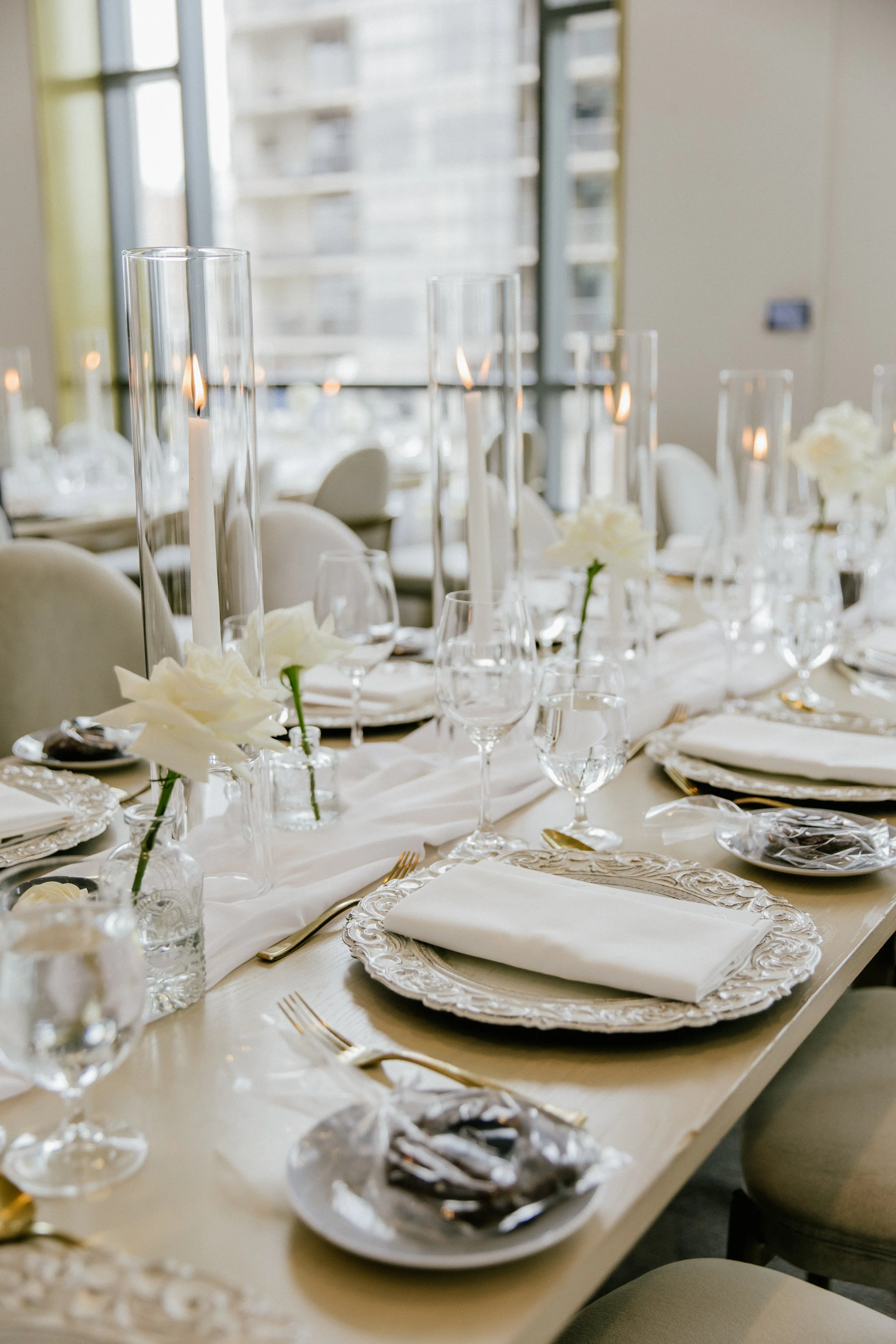 Elegant dining table set for a formal event with white flowers, candles, glassware, and gold utensils in a bright room.