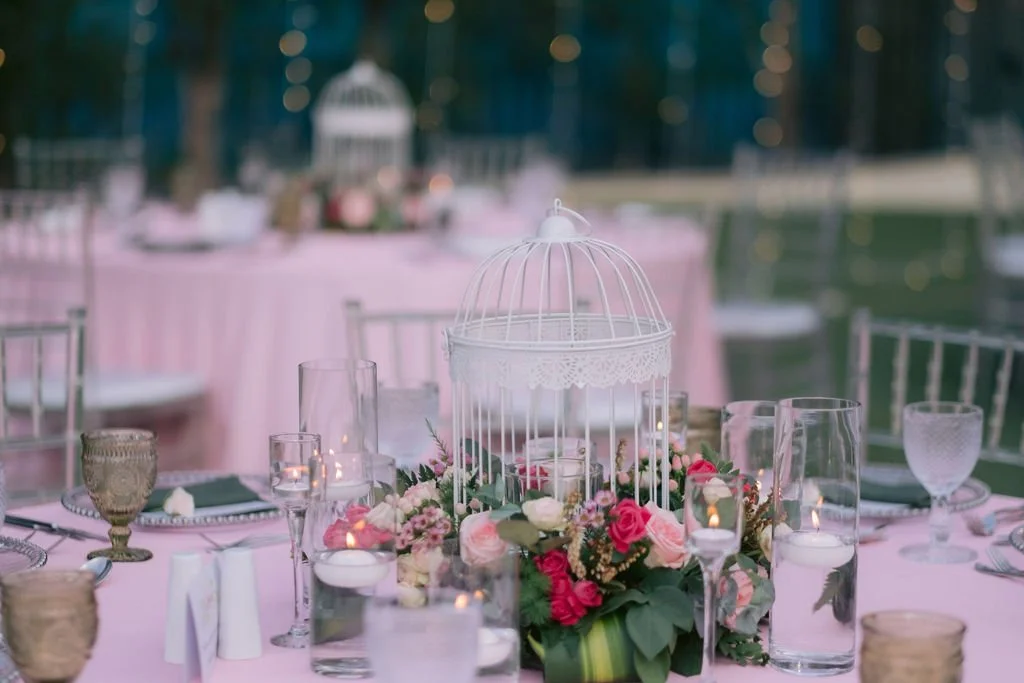 Wedding reception table decorated with pink tablecloths, floral arrangements, candles, glassware, and a white birdcage centerpiece.