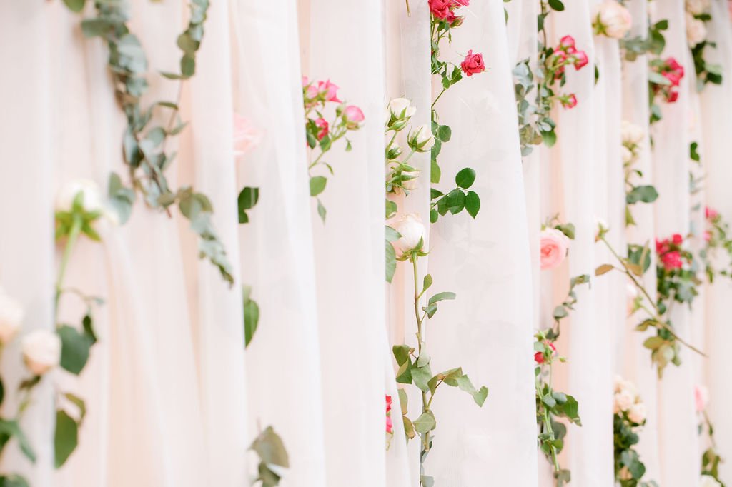 Close-up of a floral curtain with pink and white roses and green leaves behind a sheer white fabric.