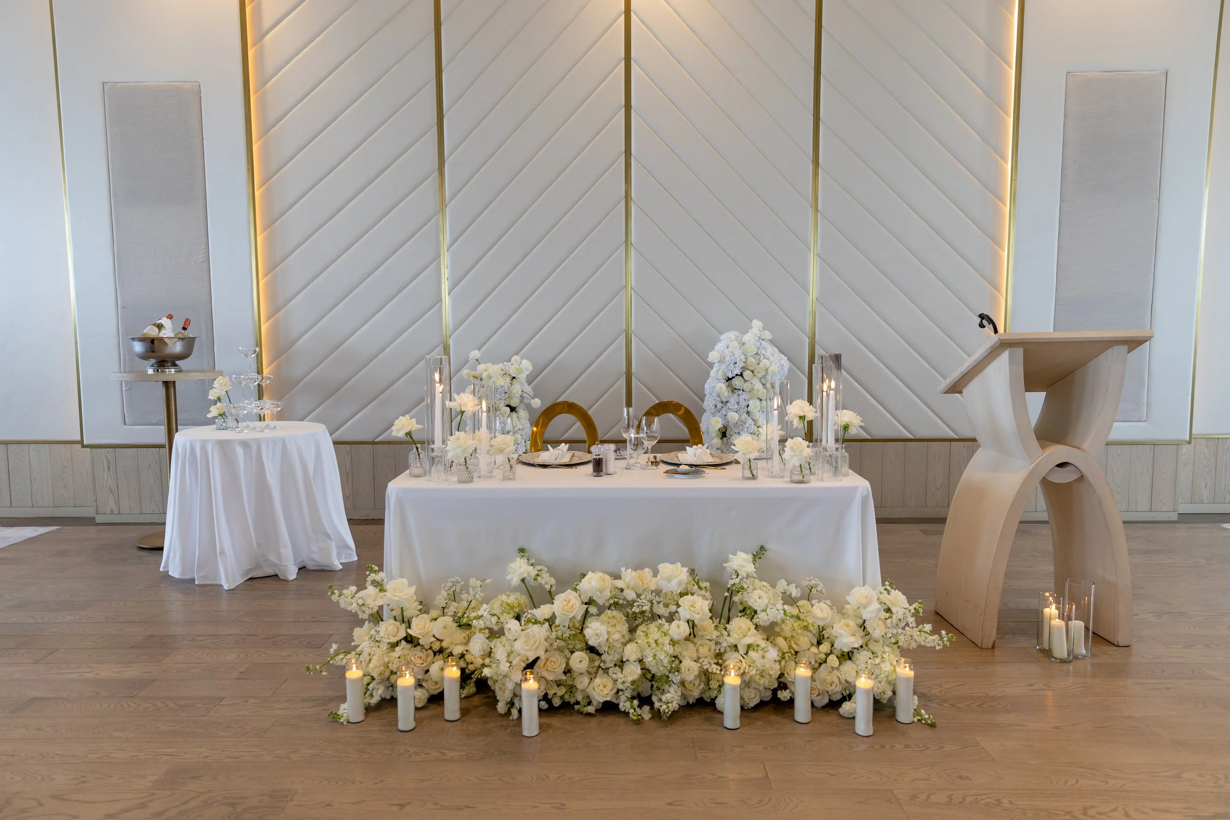 Wedding reception table decorated with white flowers, candles, and gold accents in an elegant room.