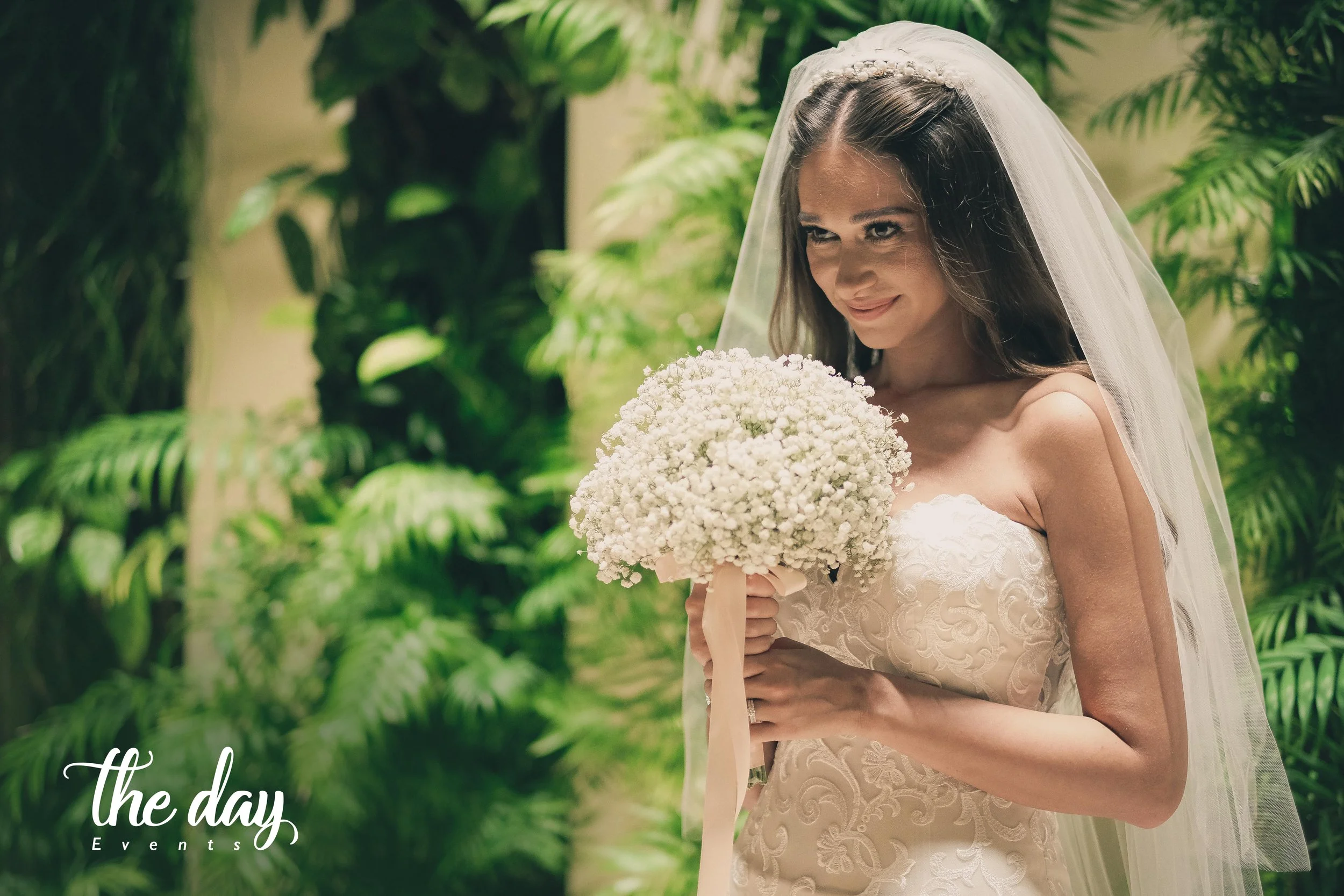 A bride in a white wedding gown with a veil holding a bouquet of white flowers, standing in greenery.