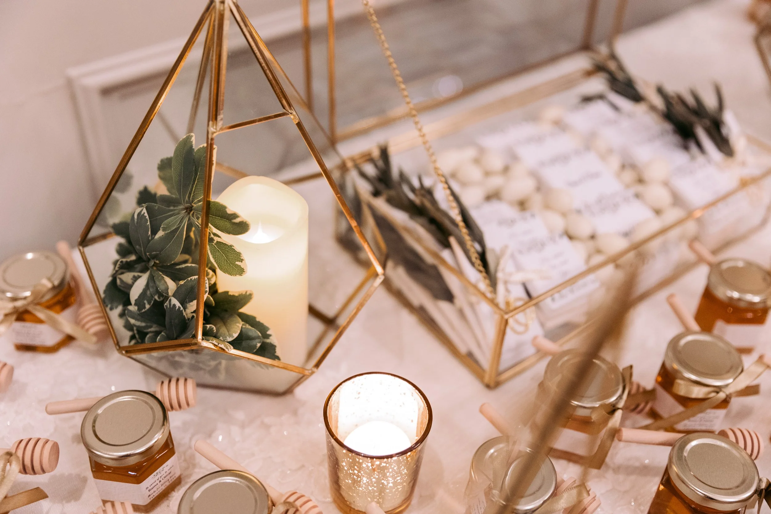 Decorative display with glass geometric terrarium containing a candle and greenery, jars of honey with dip sticks, and a glass container filled with small wrapped items, all arranged on a table.