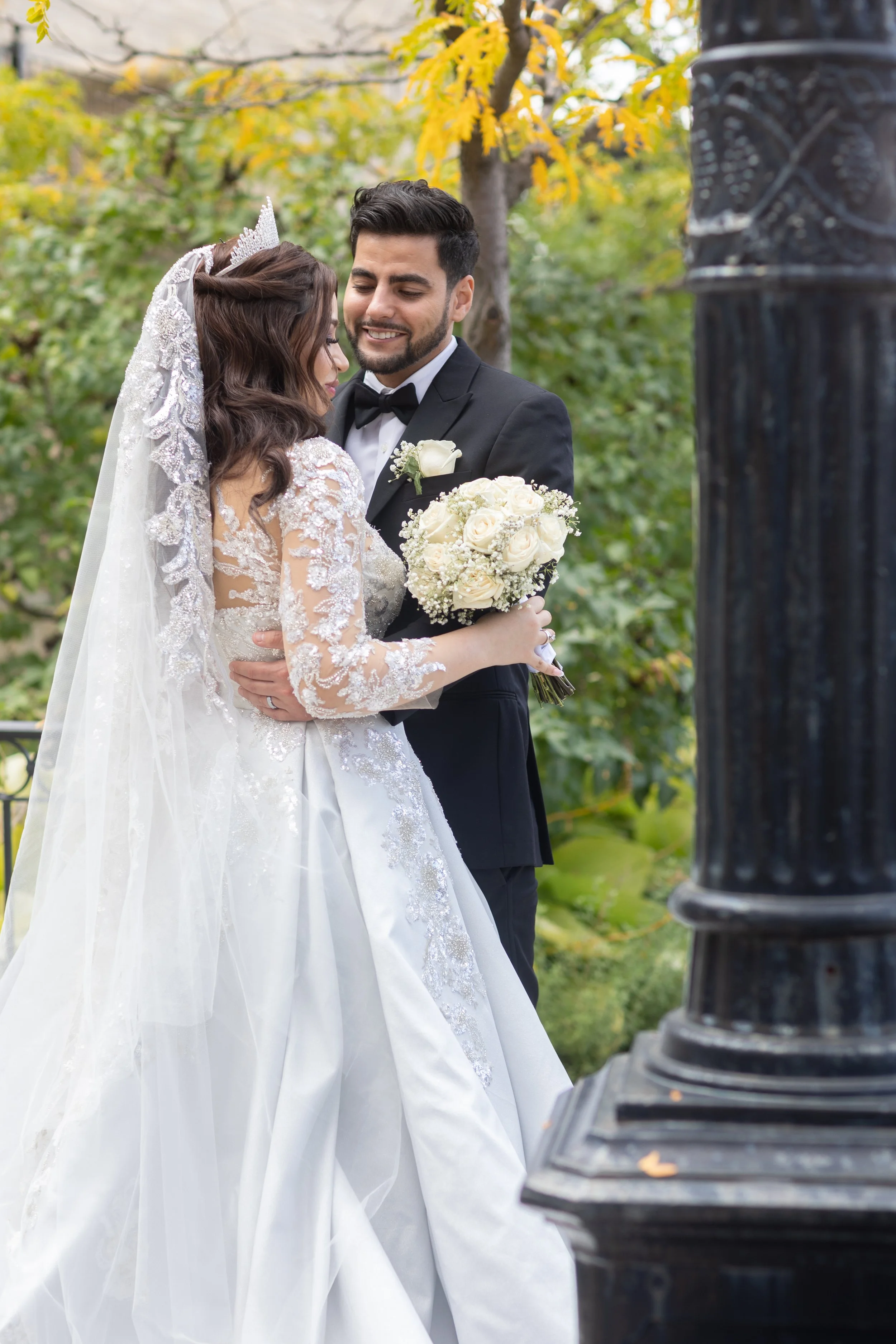 A bride and groom embrace outdoors during their wedding, with the bride wearing a white lace wedding dress and veil, holding a bouquet of white roses, and the groom in a black tuxedo with a bow tie. They are surrounded by green and yellow autumn tree