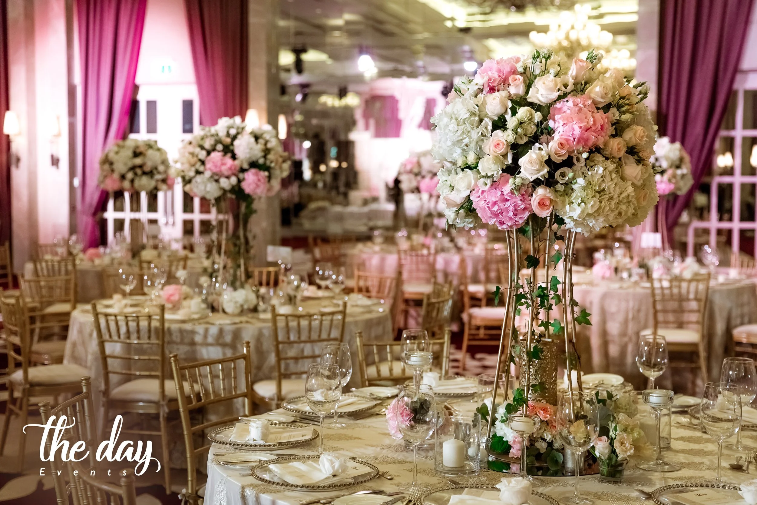 Elegant event reception with round tables decorated with large pink and white floral centerpieces, beige tablecloths, and gold chairs in a decorated venue with pink drapes and chandeliers.