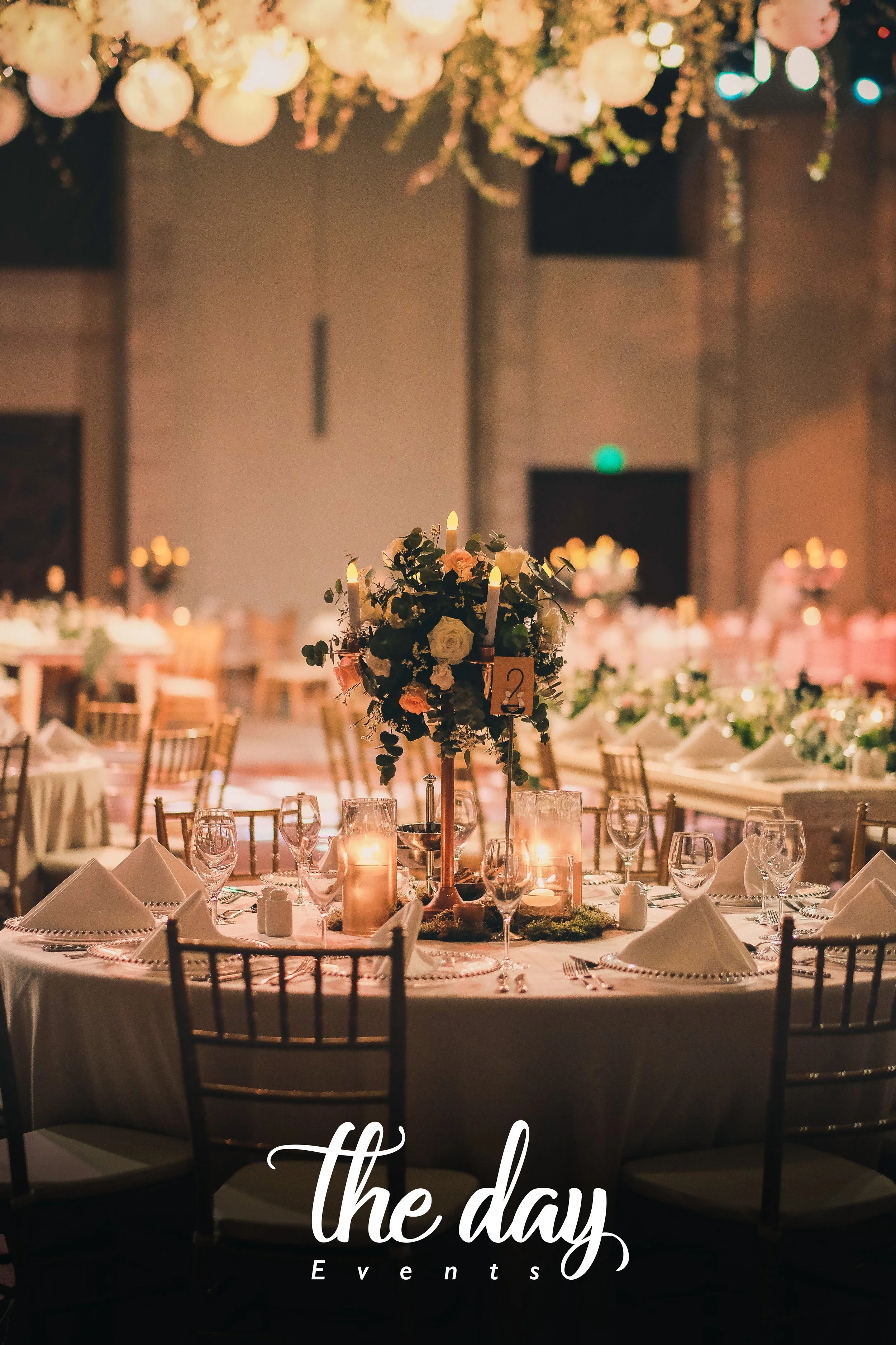 Elegant banquet table setup with candles, floral centerpiece, wine glasses, folded napkins, and table number card in a dimly lit event hall decorated with hanging floral arrangements.