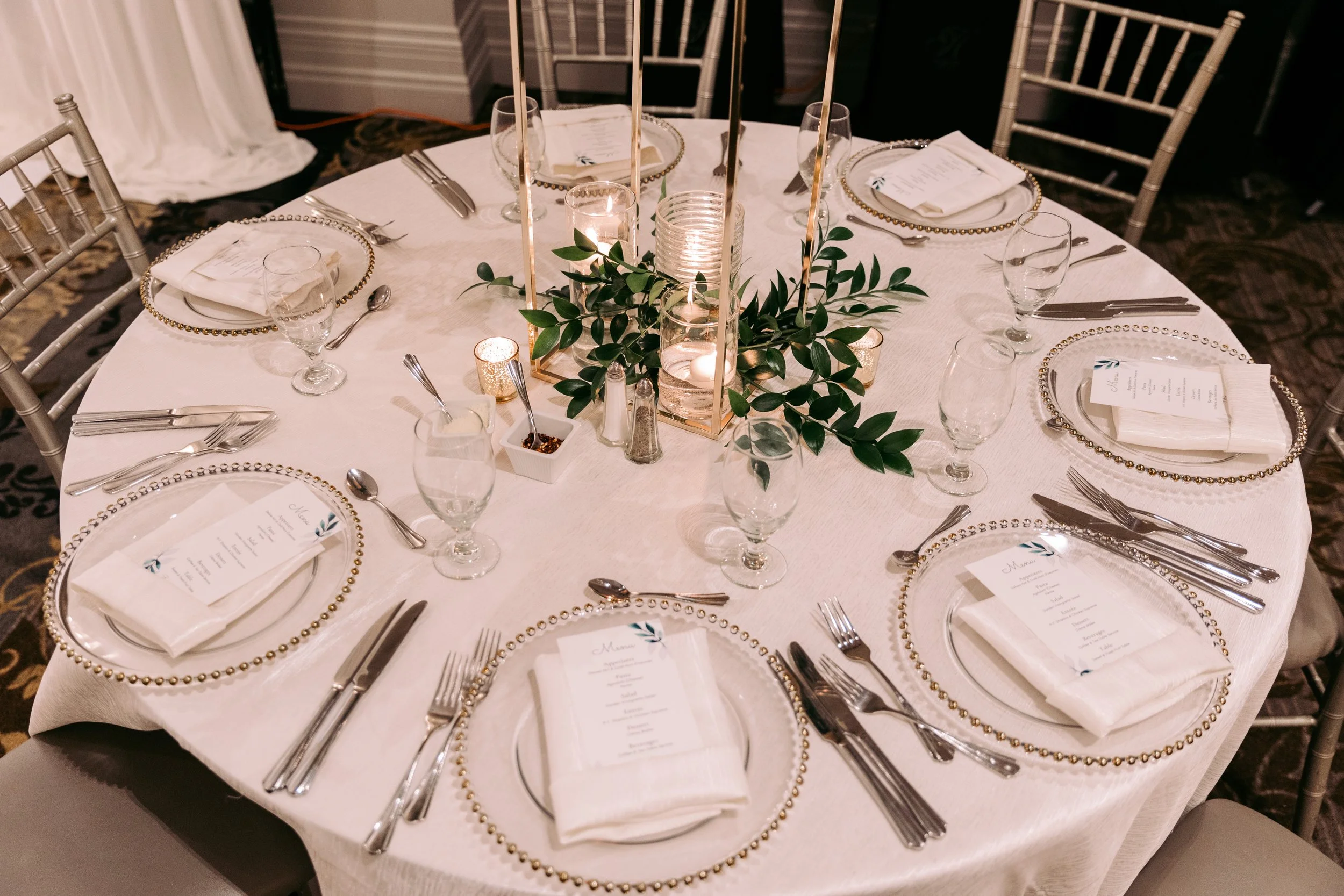 Round banquet table set for an elegant event with white tablecloth, gold beaded chargers, folded napkins, printed menus, wine glasses, and silverware. A centerpiece features candles, green foliage, and gold accents.