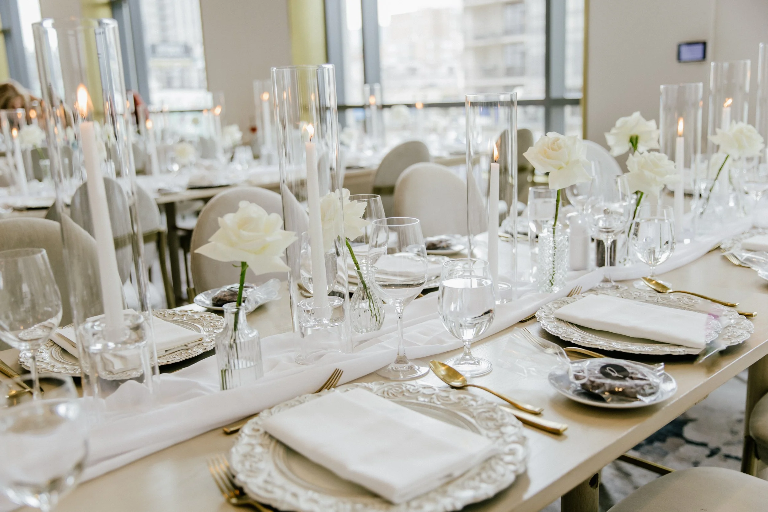Elegant dining table set with white floral arrangements, glassware, candles, and gold utensils in a bright room with large windows.