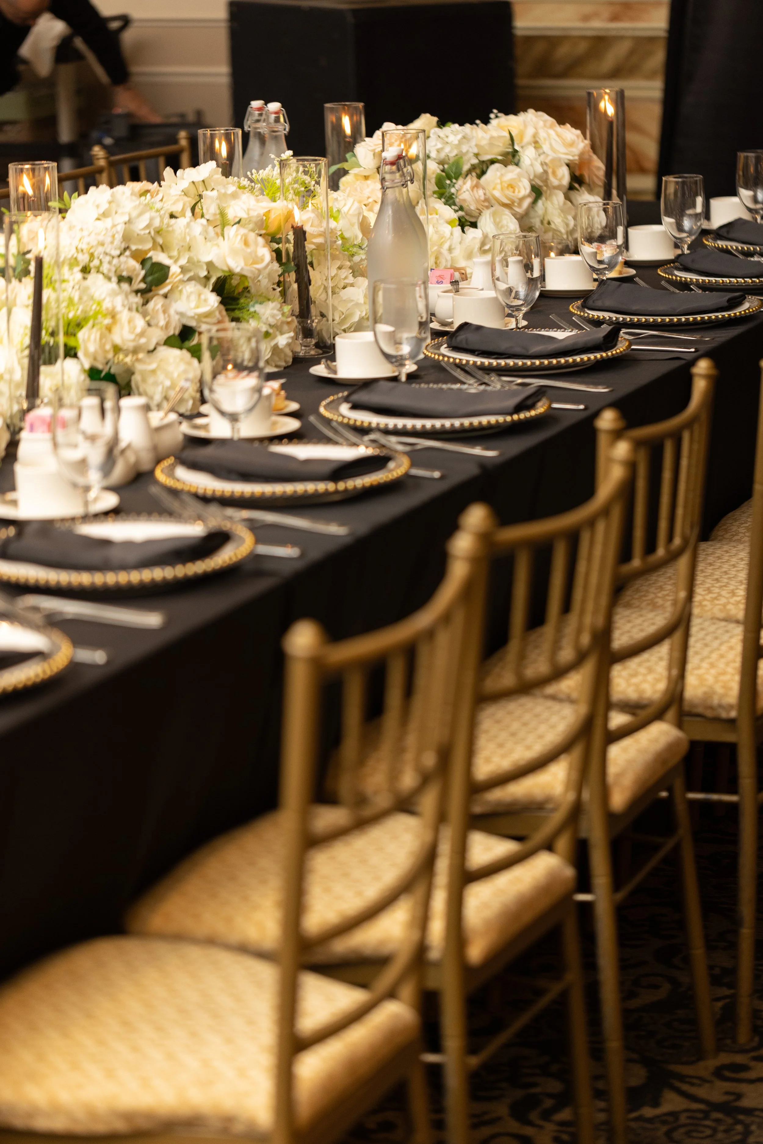 A banquet table set for an event with black tablecloth, gold chairs, white floral centerpieces, black and gold plates, black napkins, clear glassware, and candles.
