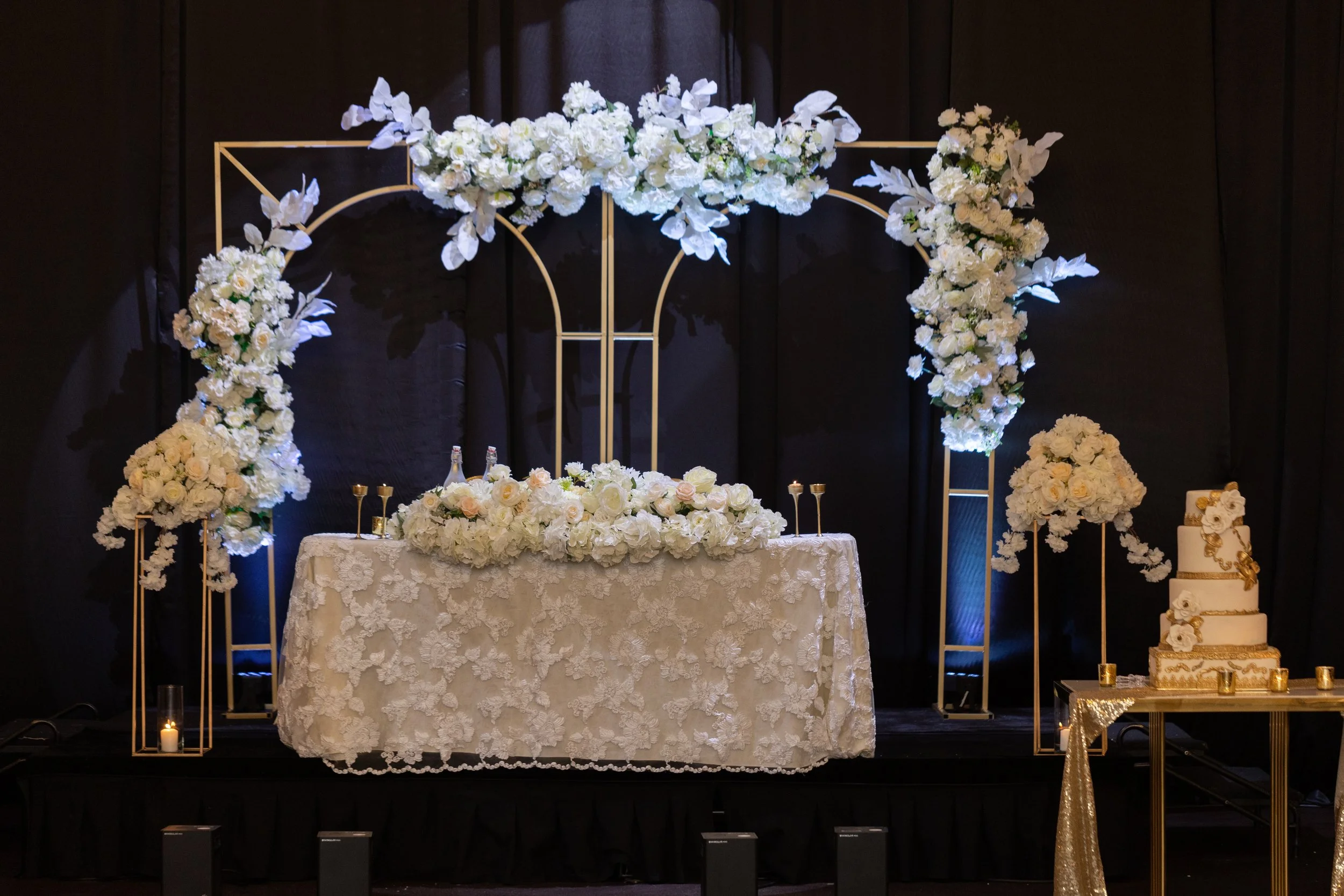 Wedding stage decorated with white flowers, gold accents, candles, and a tiered wedding cake with white and gold decorations.