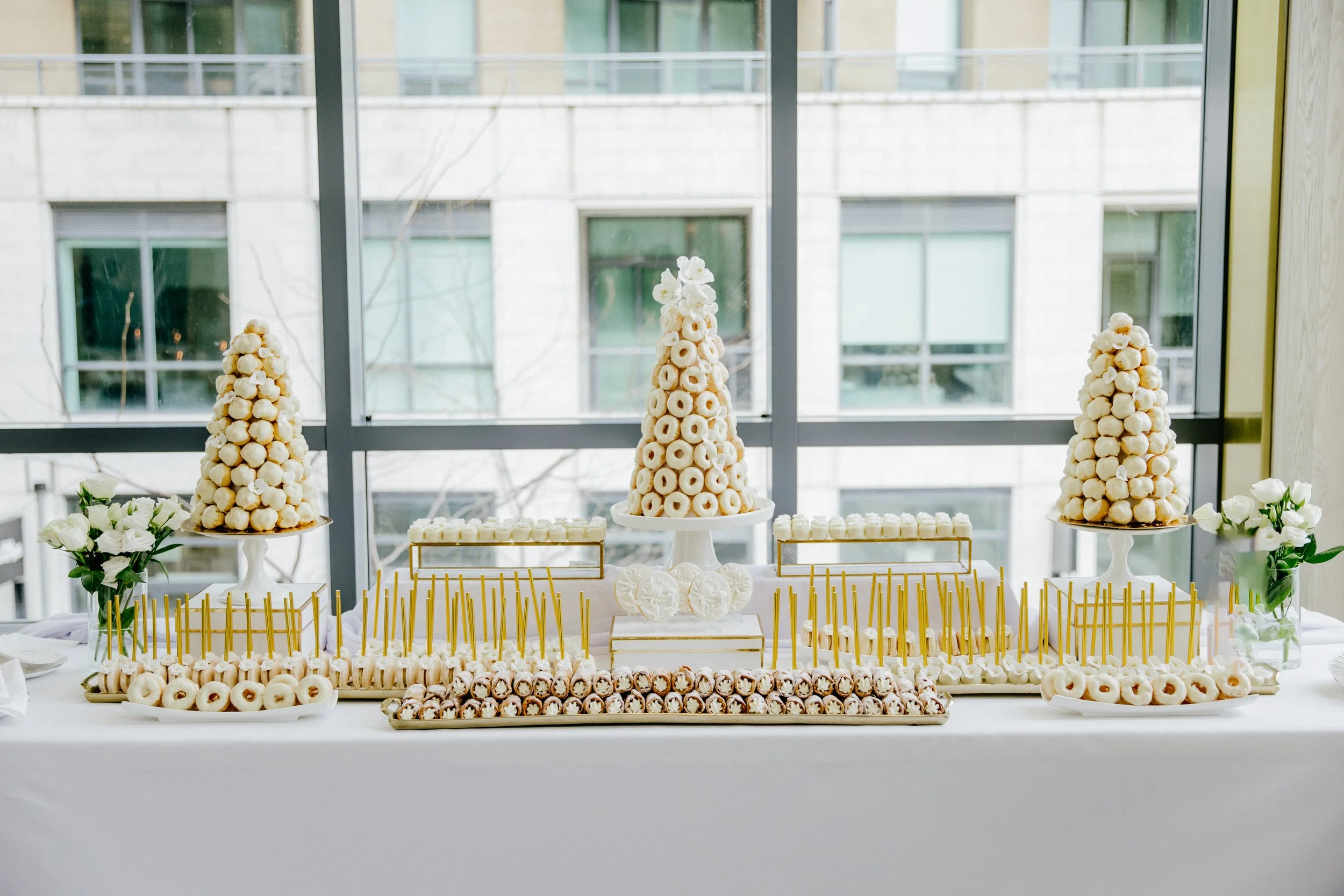 Elegant dessert table with white and gold treats, including a central cone of ring-shaped cookies, surrounded by plates of white donuts, cake pops, and other confections, with white flowers on either side, set against large windows in the background.