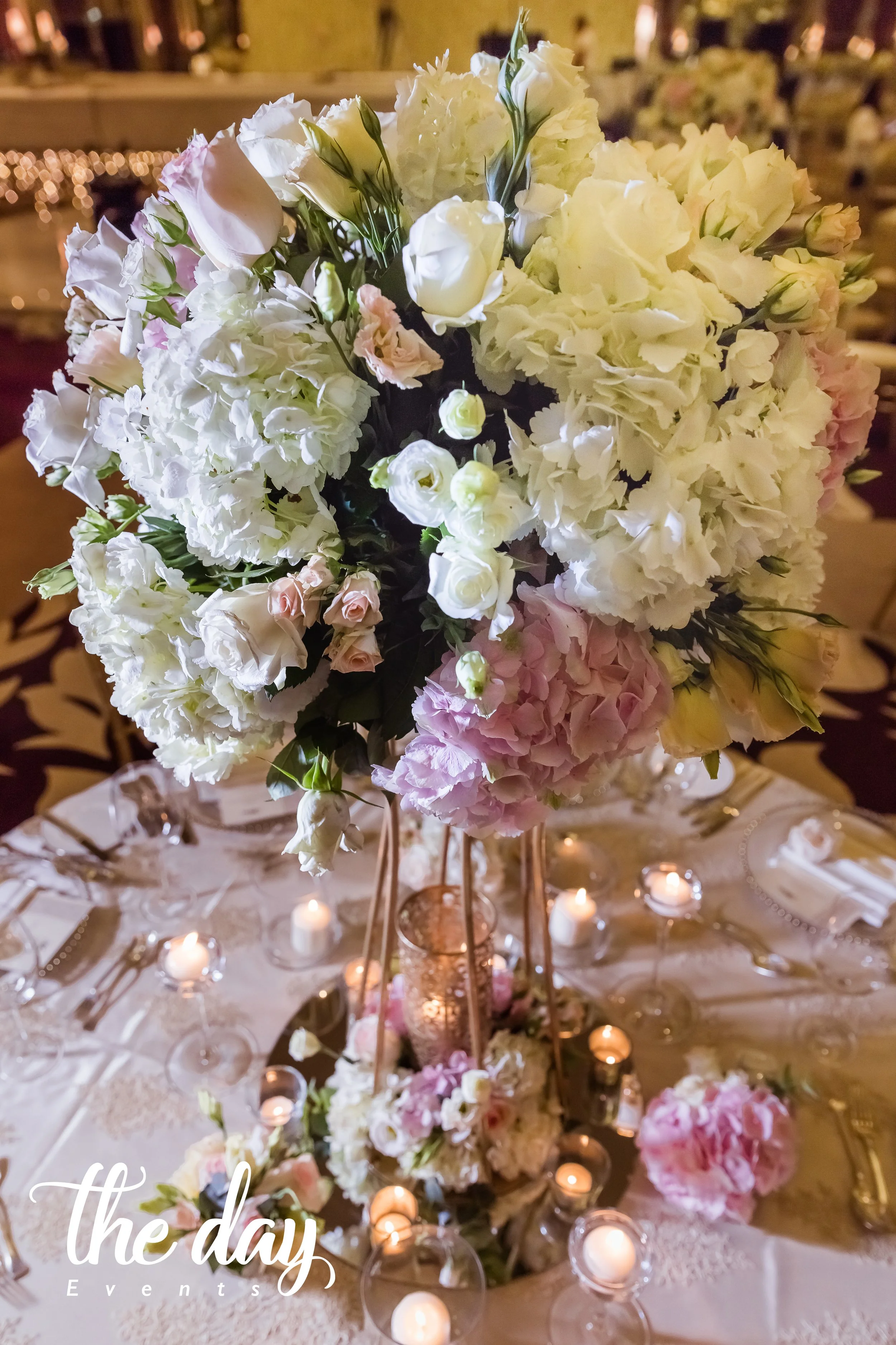 A tall floral centerpiece with white and pink flowers on a banquet table decorated with candles and elegant tableware at an event.